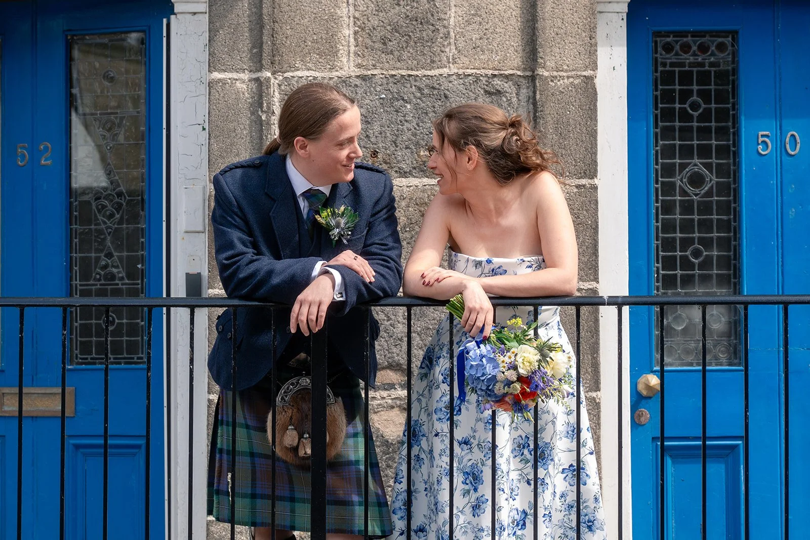 A man and woman smile at each other on a balcony outside a blue door and window. The man wears a dark blue suit and kilt, and the woman wears a white floral dress holding a bouquet of flowers.