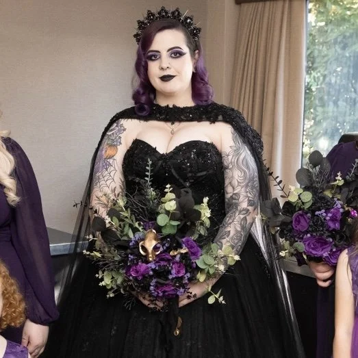 A woman with purple hair and makeup dressed in a black gothic gown with lace sleeves, holding a bouquet of purple and green flowers.
