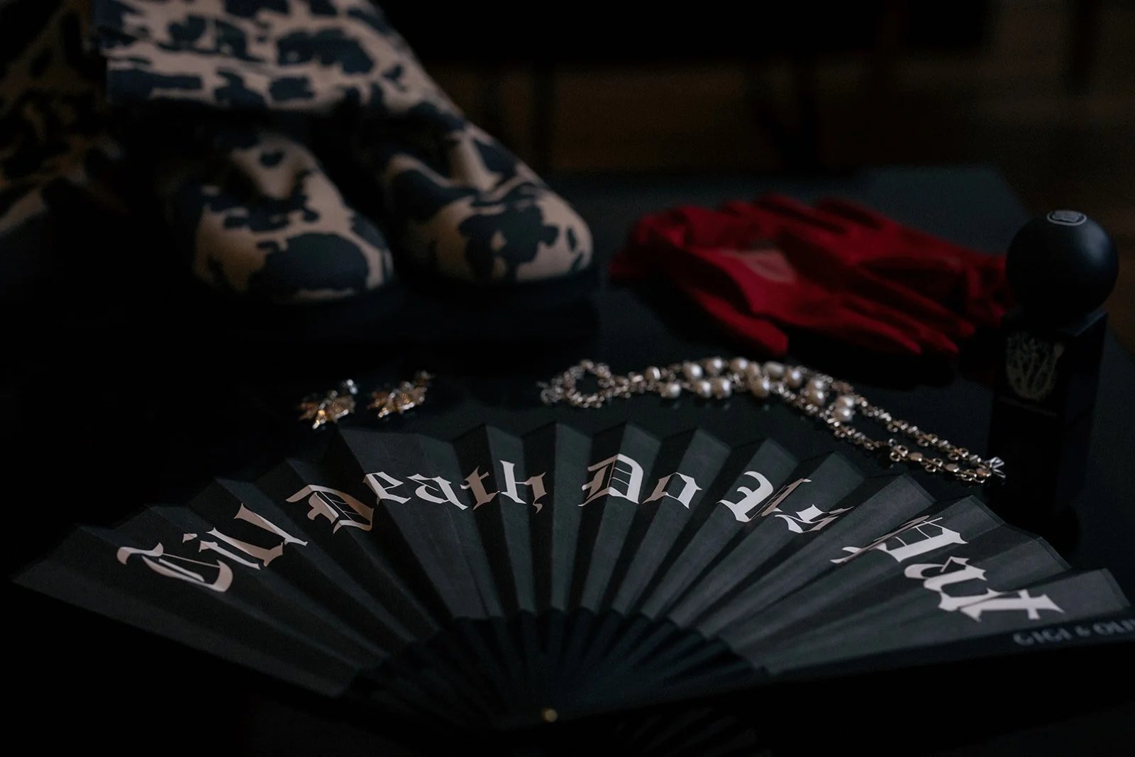 Goth-themed accessories and items on a dark surface, including a black paper fan with white gothic text, pearl jewelry, a red silk scarf, camouflage slippers, and a small black container with a lid.