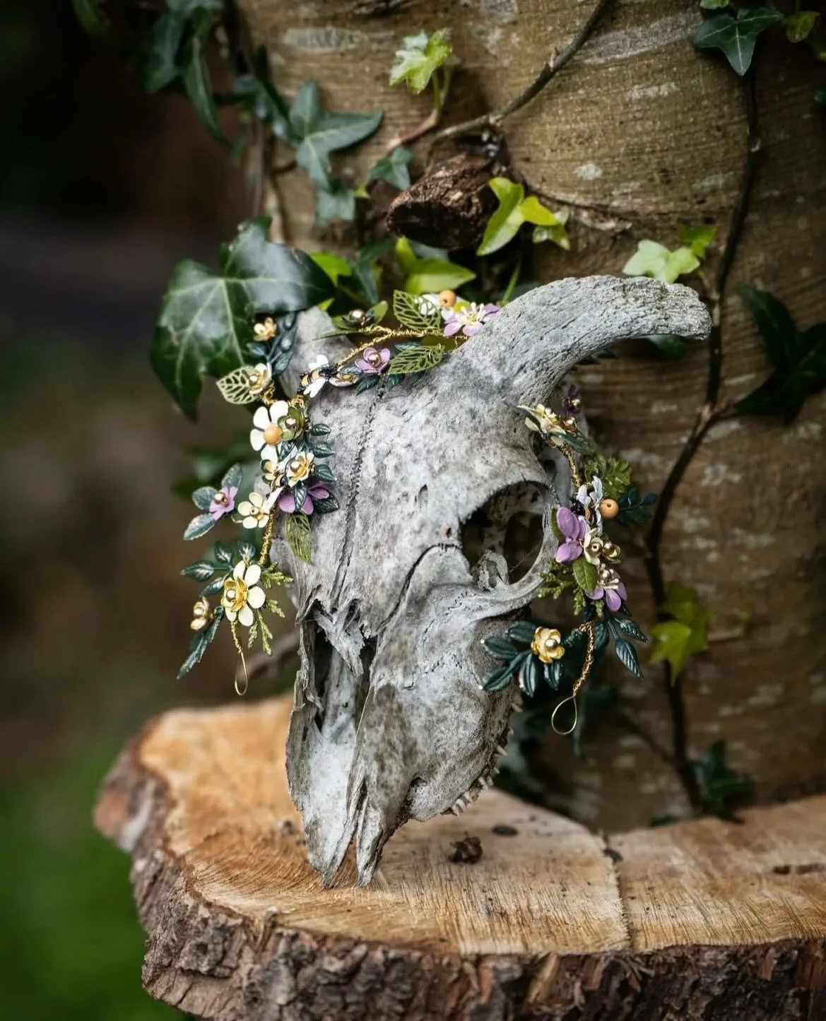 Decorative goat skull with artificial flowers and leaves, placed on a wooden surface with a wooden wall and ivy in the background.