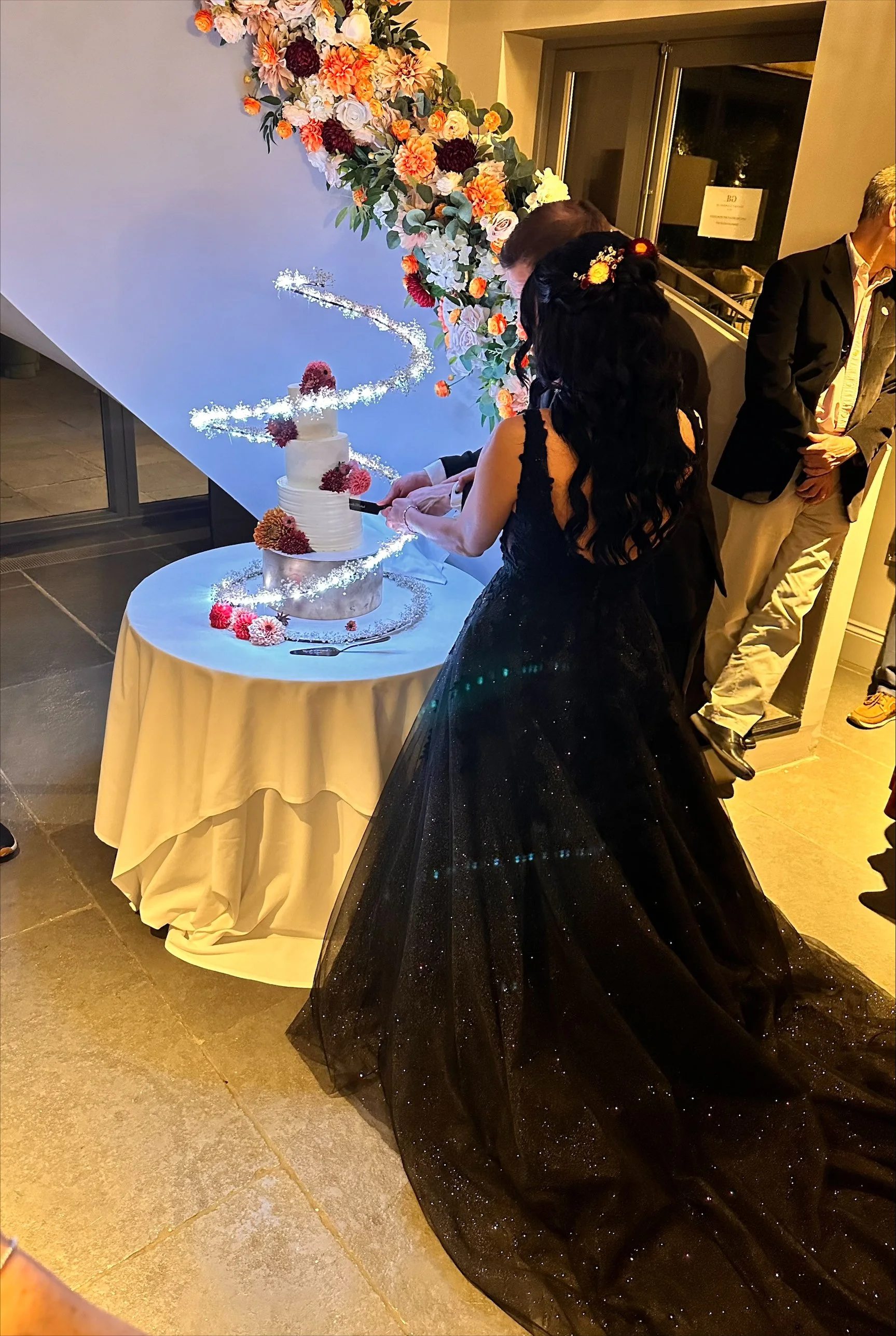A woman in a black, starry gown is cutting a tiered white wedding cake decorated with flowers, at a wedding reception. She has dark, wavy hair with floral accessories. There is a floral arch and other guests in the background.