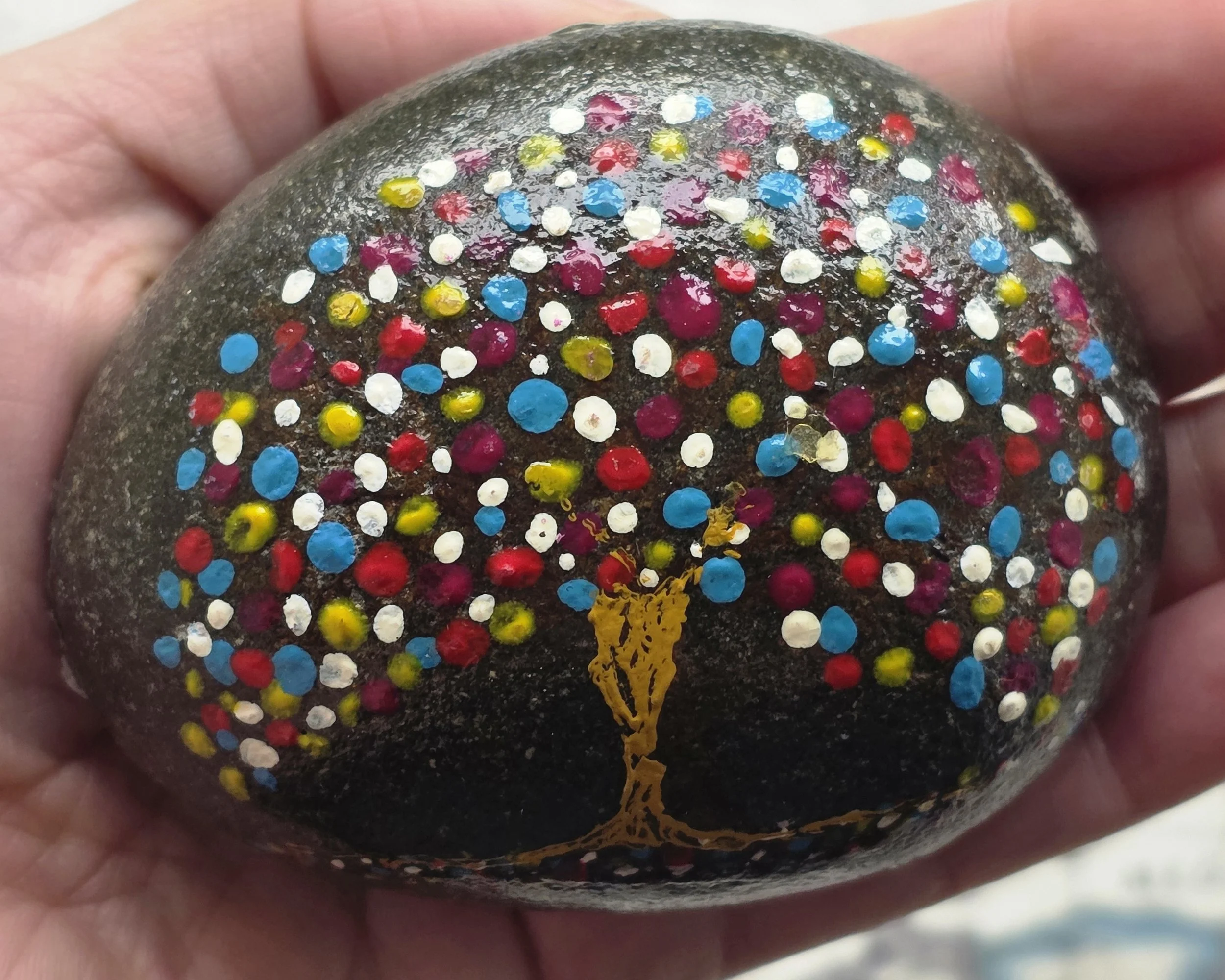 A hand holding a black rock decorated with colorful dots of paint in blue, white, pink, red, and yellow, and a gold-colored tree design painted on it.