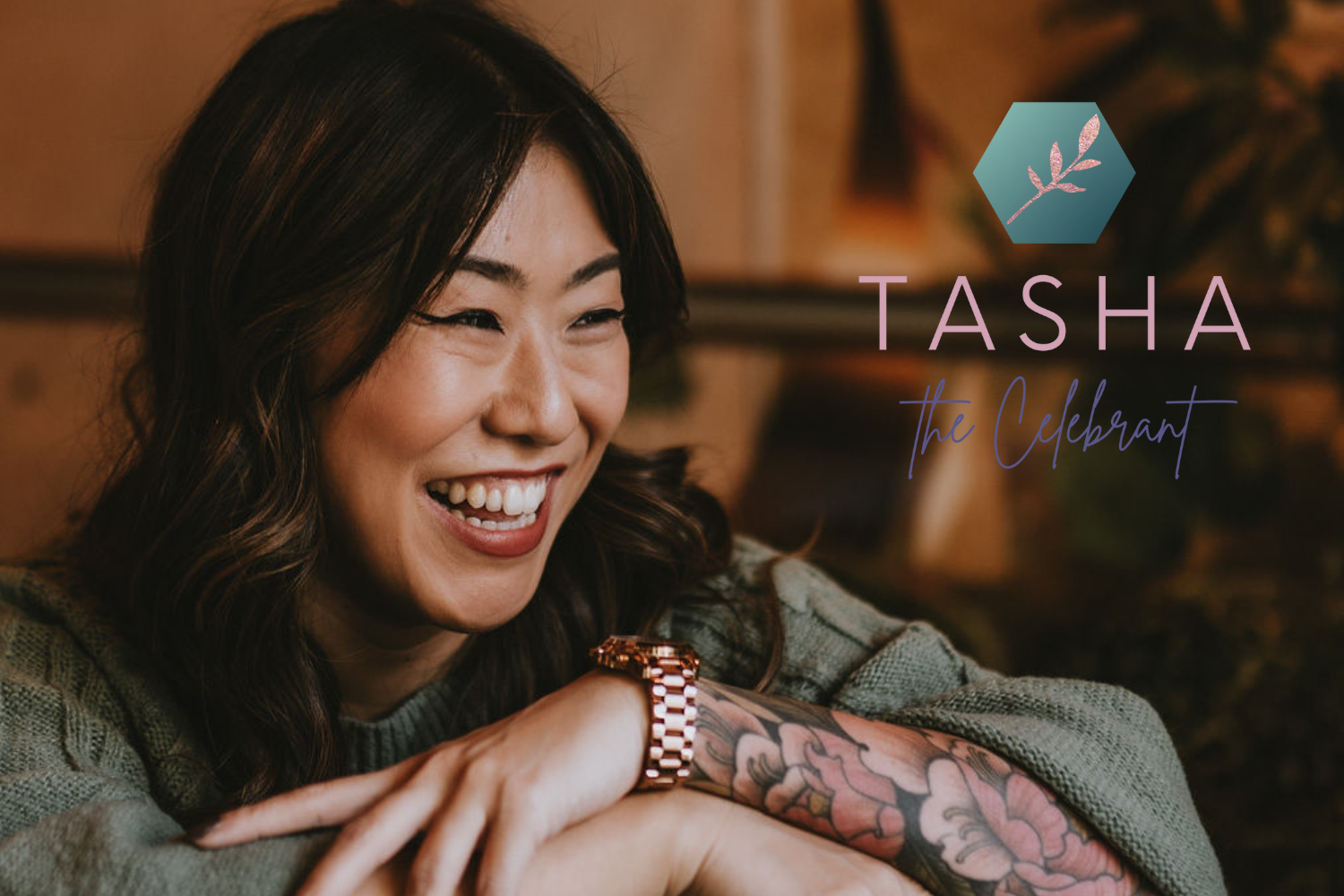 A woman with dark hair smiling and showing teeth, wearing a green sweater and a gold watch, with a tattooed arm resting on a surface, in a cozy indoor setting with wooden decor and plant in the background, alongside overlay text that reads 'Tasha: The Celebrant'.