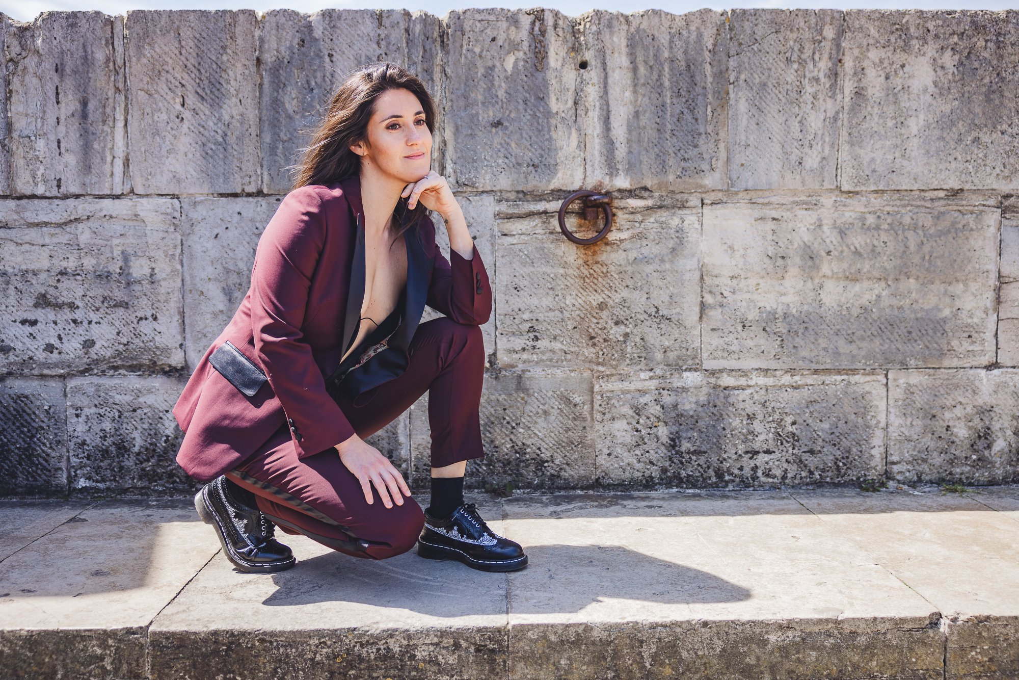 Woman in a maroon suit crouching on the sidewalk against a stone wall.