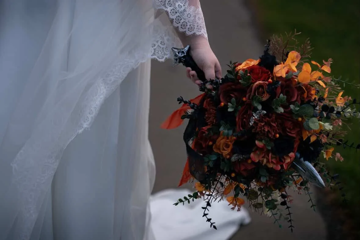 A bride in a white dress holding a vibrant bouquet of red, orange, and dark-colored flowers with greenery.