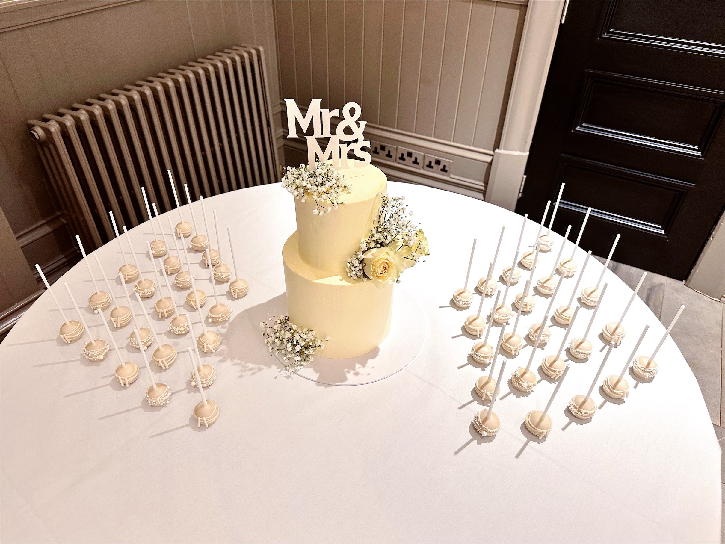 Two-tiered cream wedding cake decorated with white flowers and a 'Mr & Mrs' topper with surrounded by white cake pops on a round table covered with a white tablecloth.