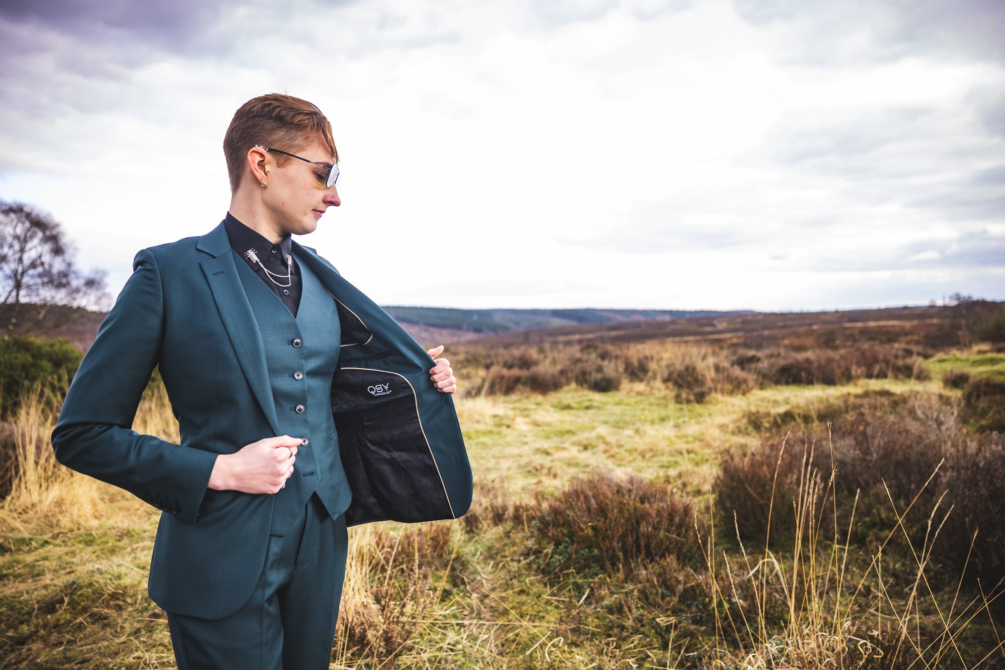 Woman in teal suit opening a black bag in a grassy, open landscape with cloudy sky.