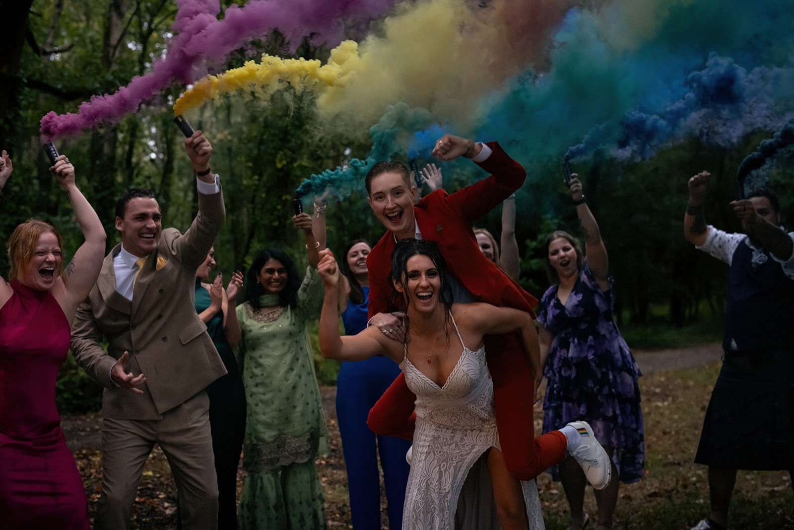 A group of people celebrating outdoors, holding colorful smoke flares, with two women in the center, one giving a piggyback ride. The people are smiling and appear joyful.