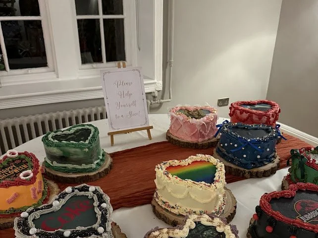 Decorative cakes shaped like heart and other designs, placed on a table with a wooden centerpiece, and a small sign that reads 'Please Help Yourself to a Slice'.