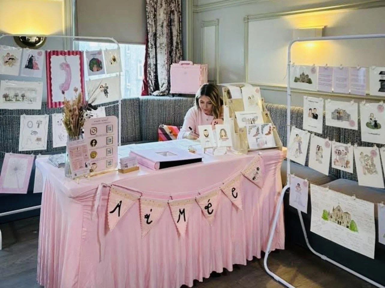 A woman at a display booth decorated with pink and white, featuring artwork, illustrations, and informational signs, with a pink banner spelling 'ÁMÉE' hanging from the front of the table.