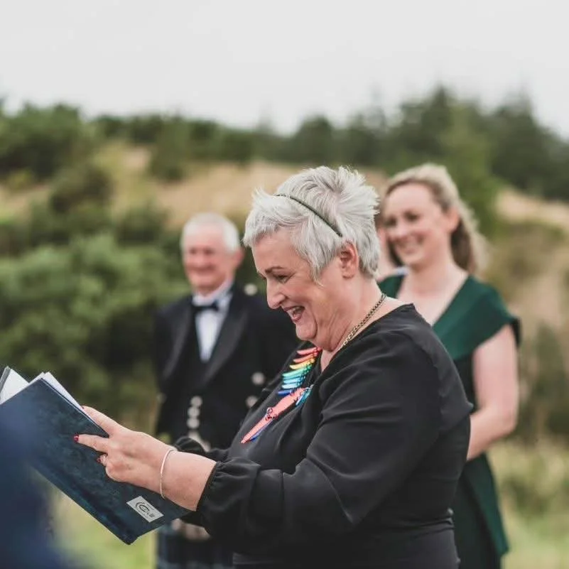 A woman with short gray hair, wearing a black outfit and necklace, is smiling and looking at a document or book she is holding. Two people, a man in a tuxedo and a woman in a green dress, stand behind her outdoors in a green, hilly area, blurred in the background.