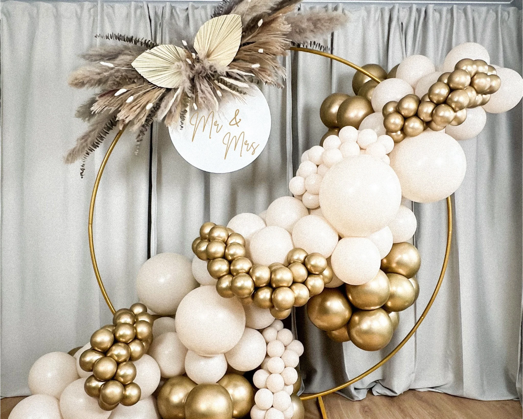 Decorative balloon arrangement with white and gold balloons, pampas grass, and a sign that reads 'Mr & Mrs' on a circular white background, arranged in a semi-circle on a gold stand.