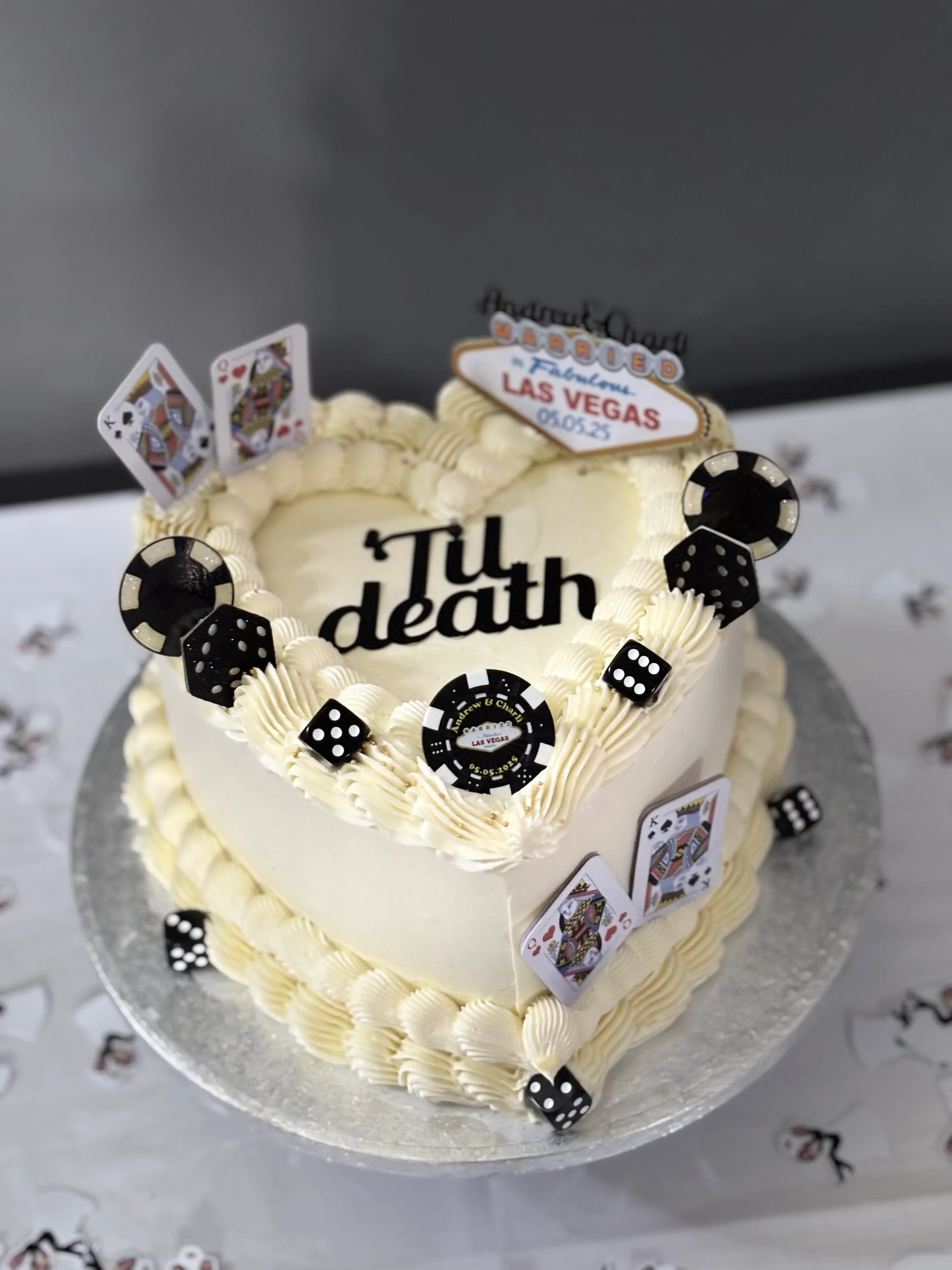 A heart-shaped celebration cake with white frosting, decorated with casino-themed edible decorations including playing cards, poker chips, and dice. The cake has a message "Till death" written on top and several themed signs, including one referencing Las Vegas and a date, 05.05.23.