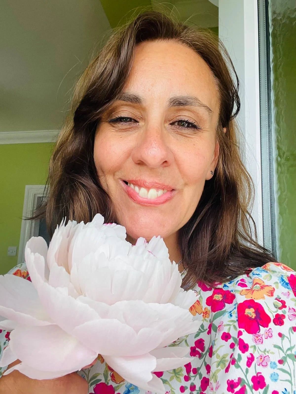 A woman smiling and holding a large white flower, standing near a window in a room with green walls.