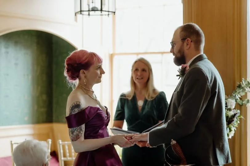 Couple exchanging vows at their wedding ceremony in a warmly lit room with an officiant, with a woman officiating and floral decorations in the background