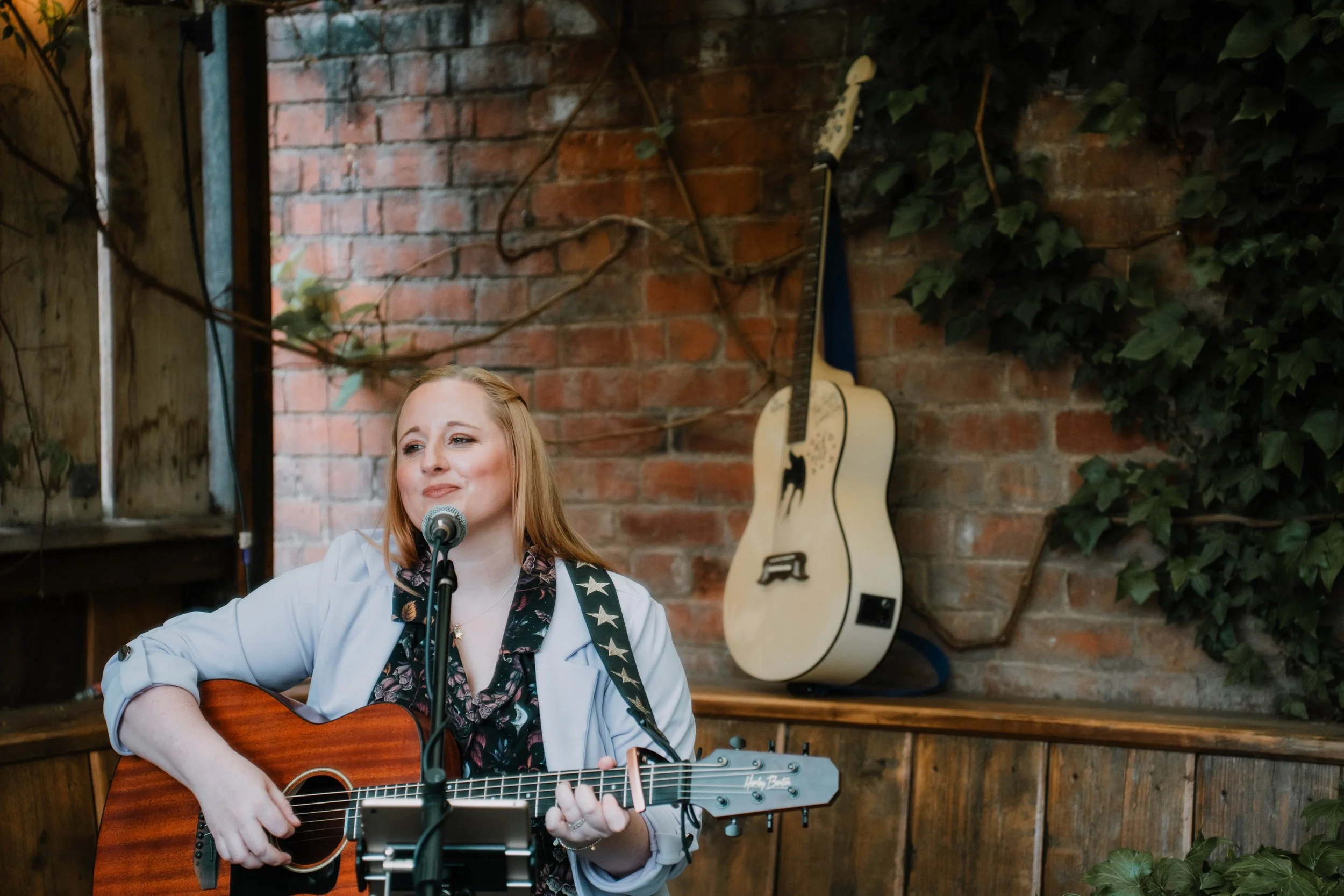 A woman with red hair playing an acoustic guitar and singing into a microphone in a cozy, rustic setting with brick walls and greenery.