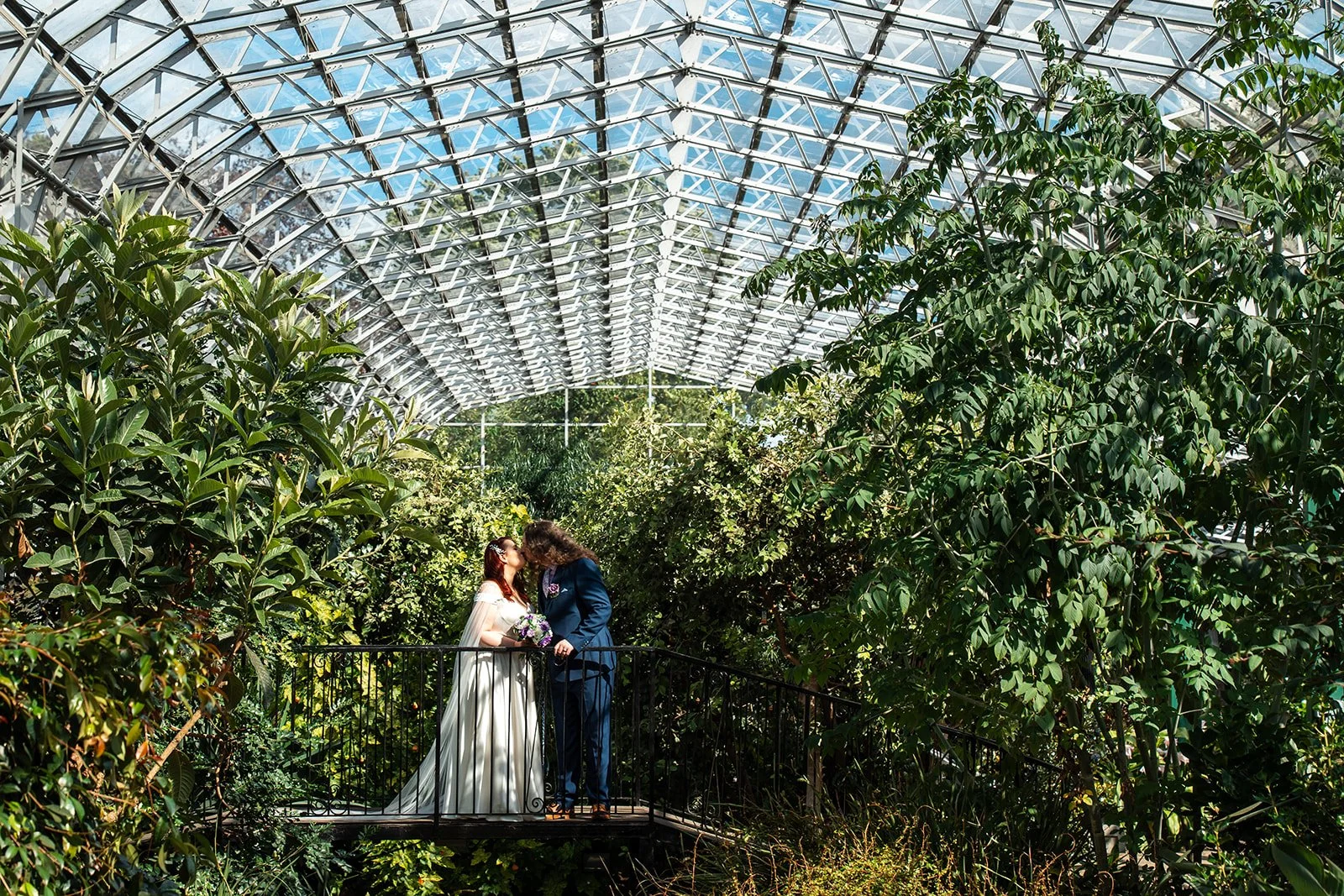 A bride and groom kissing on a small balcony surrounded by lush greenery inside a glass-domed greenhouse.