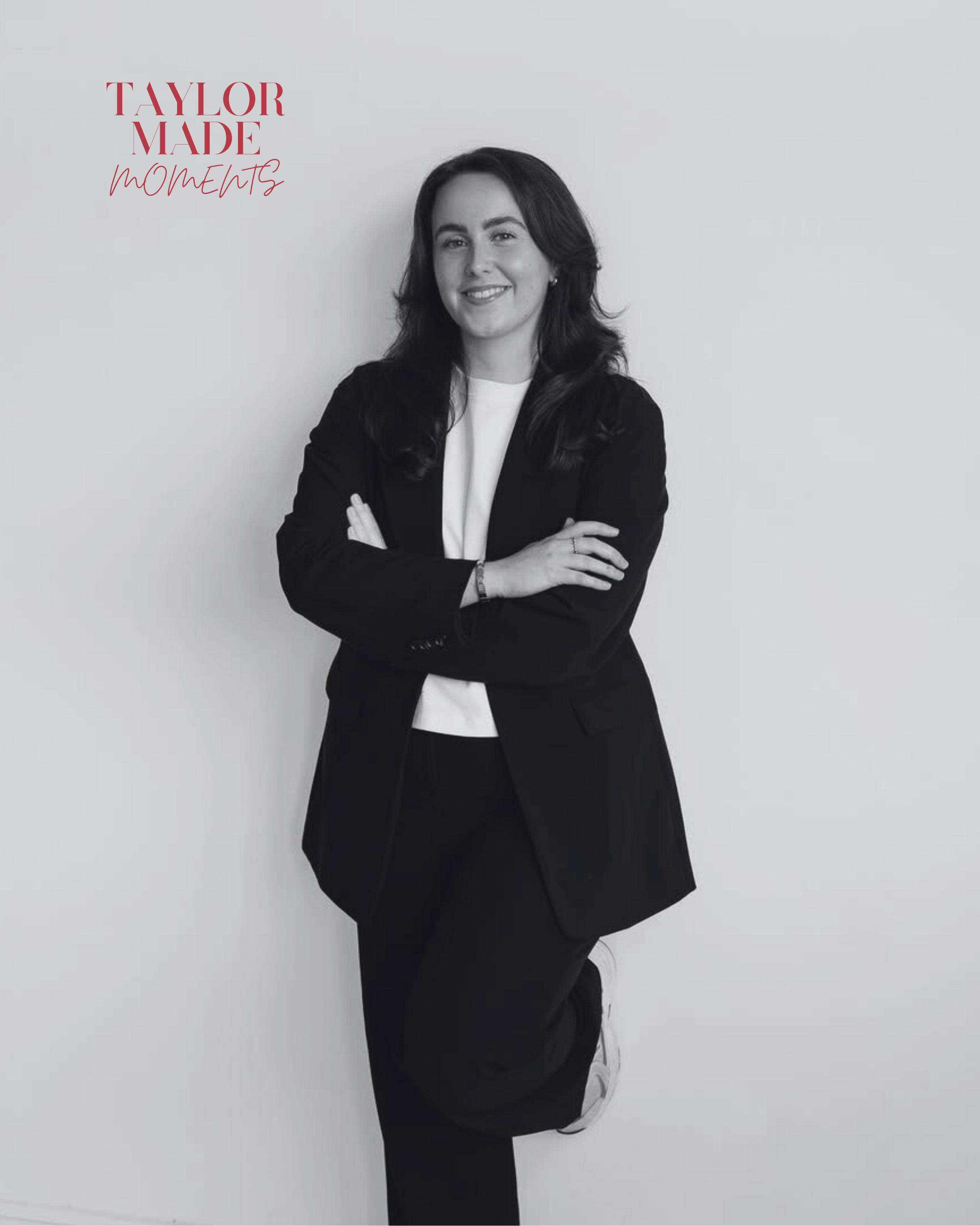 Black and white portrait of a woman with dark hair, wearing a blazer, smiling with arms crossed, standing against a plain wall. Text in the upper left corner reads 'Taylor Made Moments'.