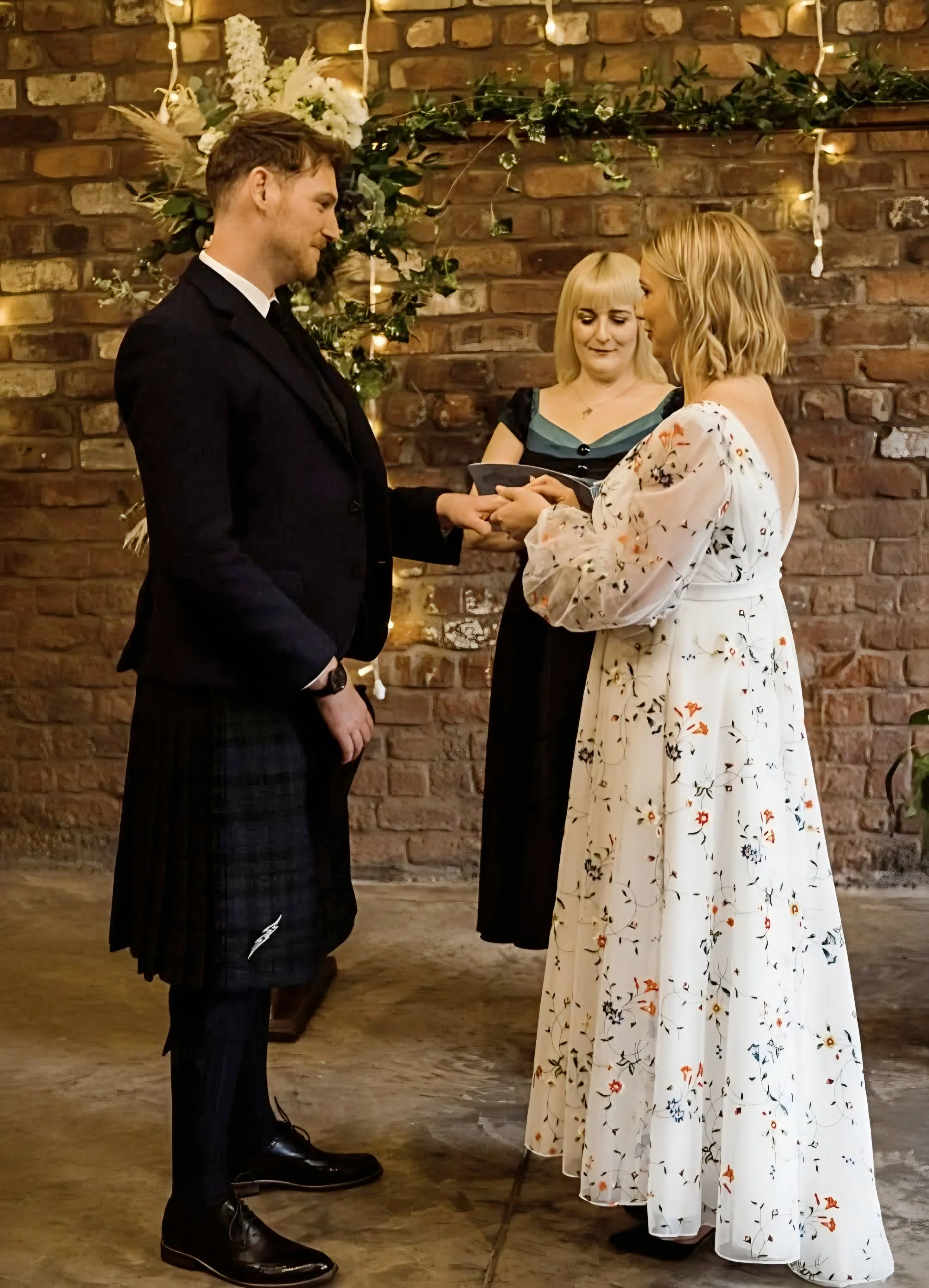 A couple exchanges vows during a wedding ceremony, with a female officiant standing behind them, in front of a decorated brick wall.