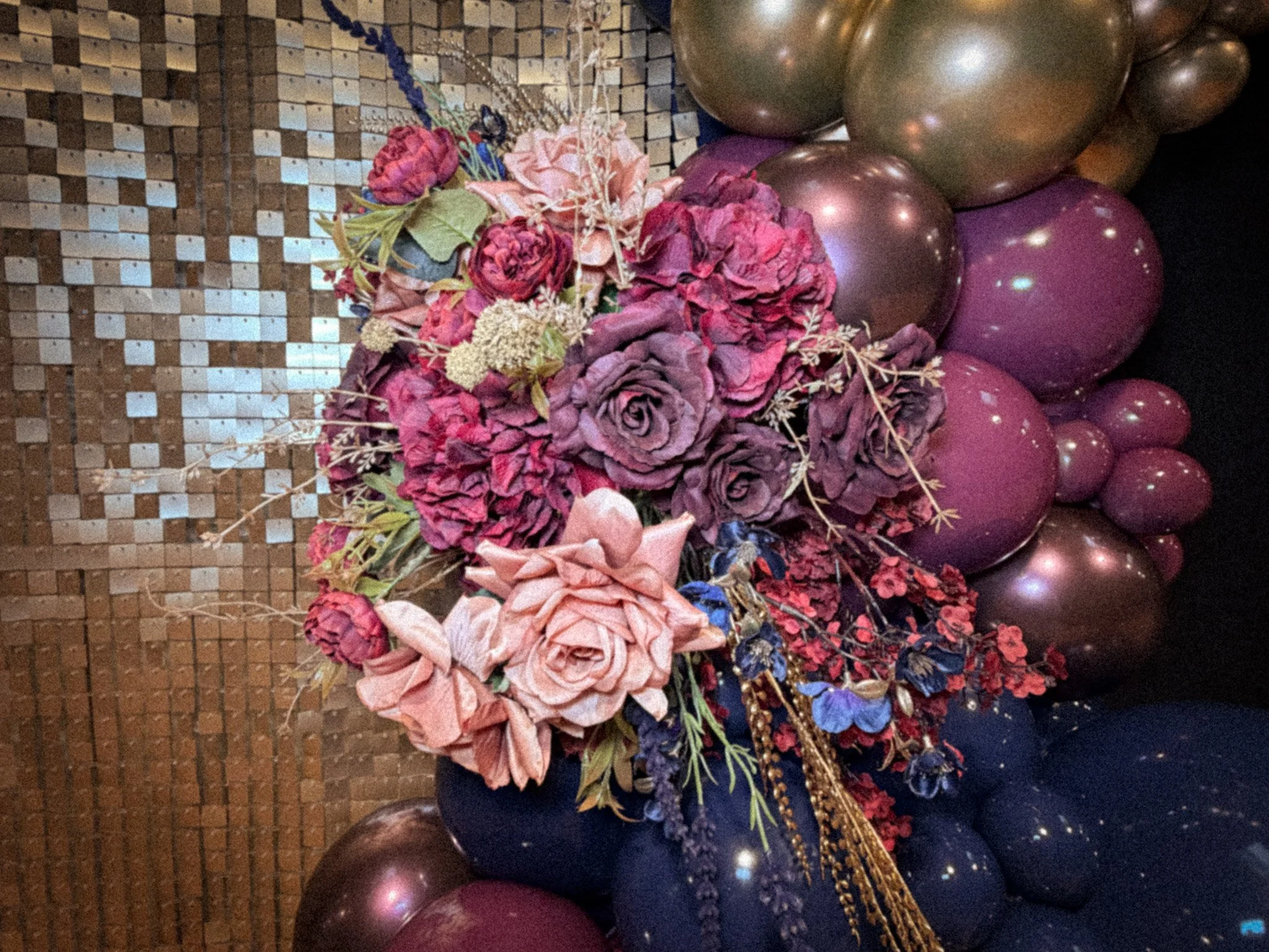 A floral arrangement featuring pink, purple, and mauve-colored flowers, surrounded by a cluster of metallic pink, purple, and bronze balloons, against a textured brown wall with small square tiles.