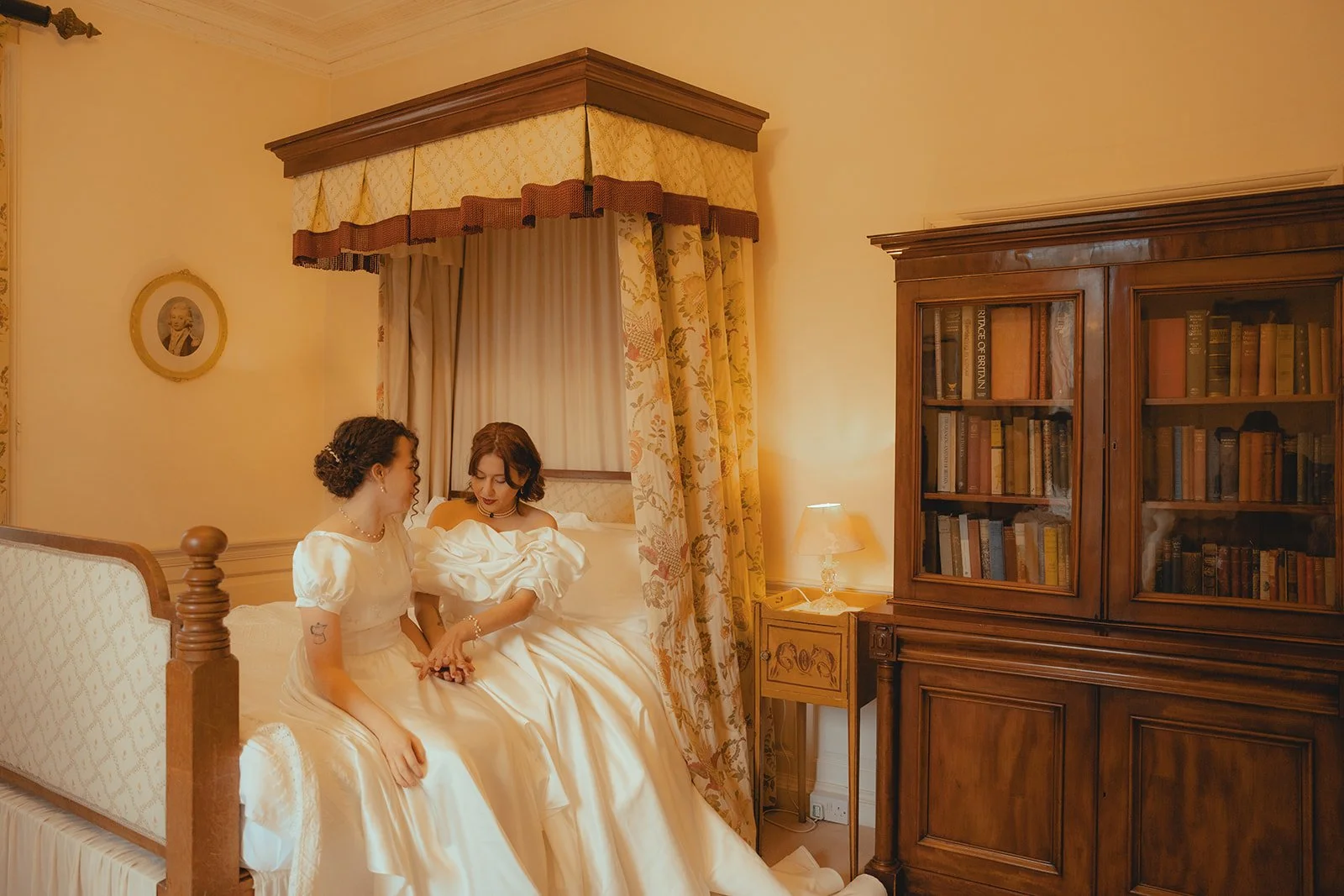 Two women in vintage style dresses sitting on a bed in a warmly lit room with antique furniture. One woman with dark curly hair, pearl necklace, and bracelet, the other with straight brown hair, holding hands and sharing a tender moment.