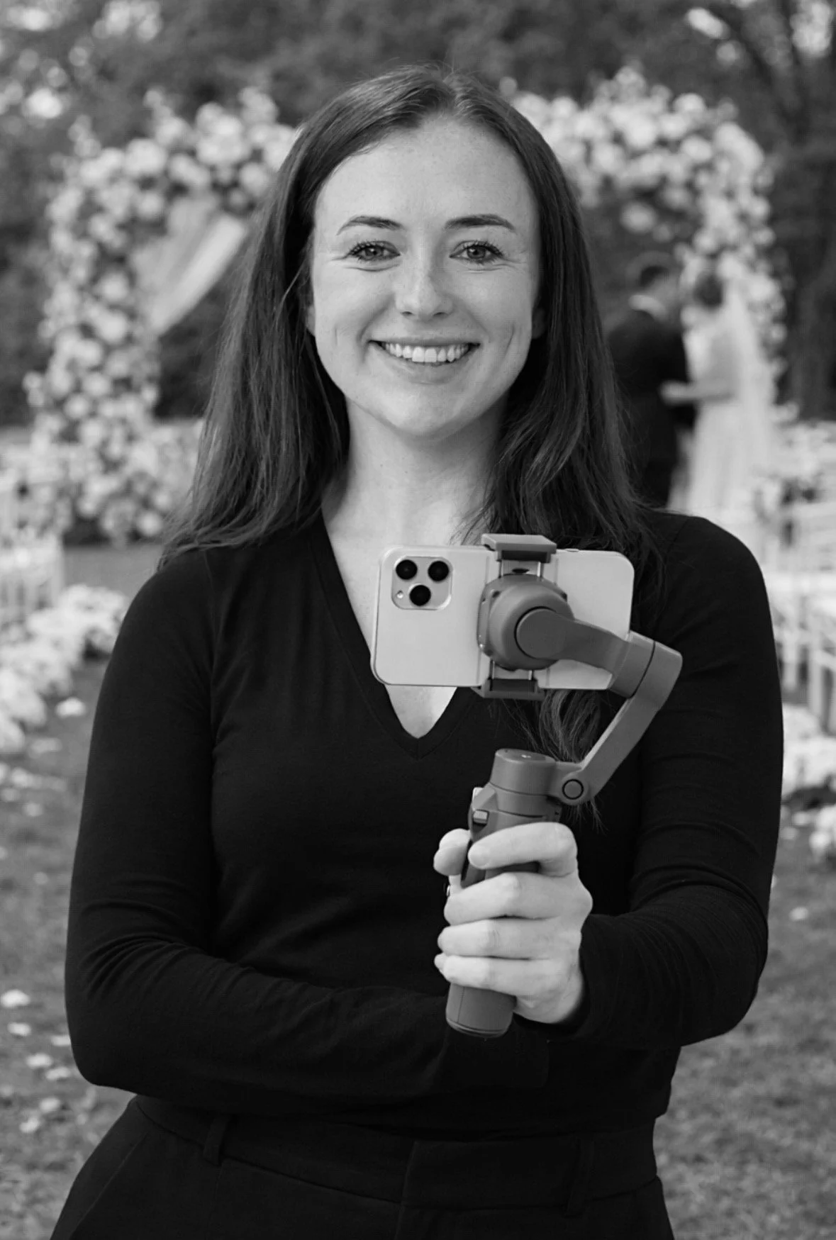 A smiling woman holding a camera mounted on a handheld stabilizer or gimbal, taking a selfie at an outdoor wedding or event, with a floral arch and other people in the background.