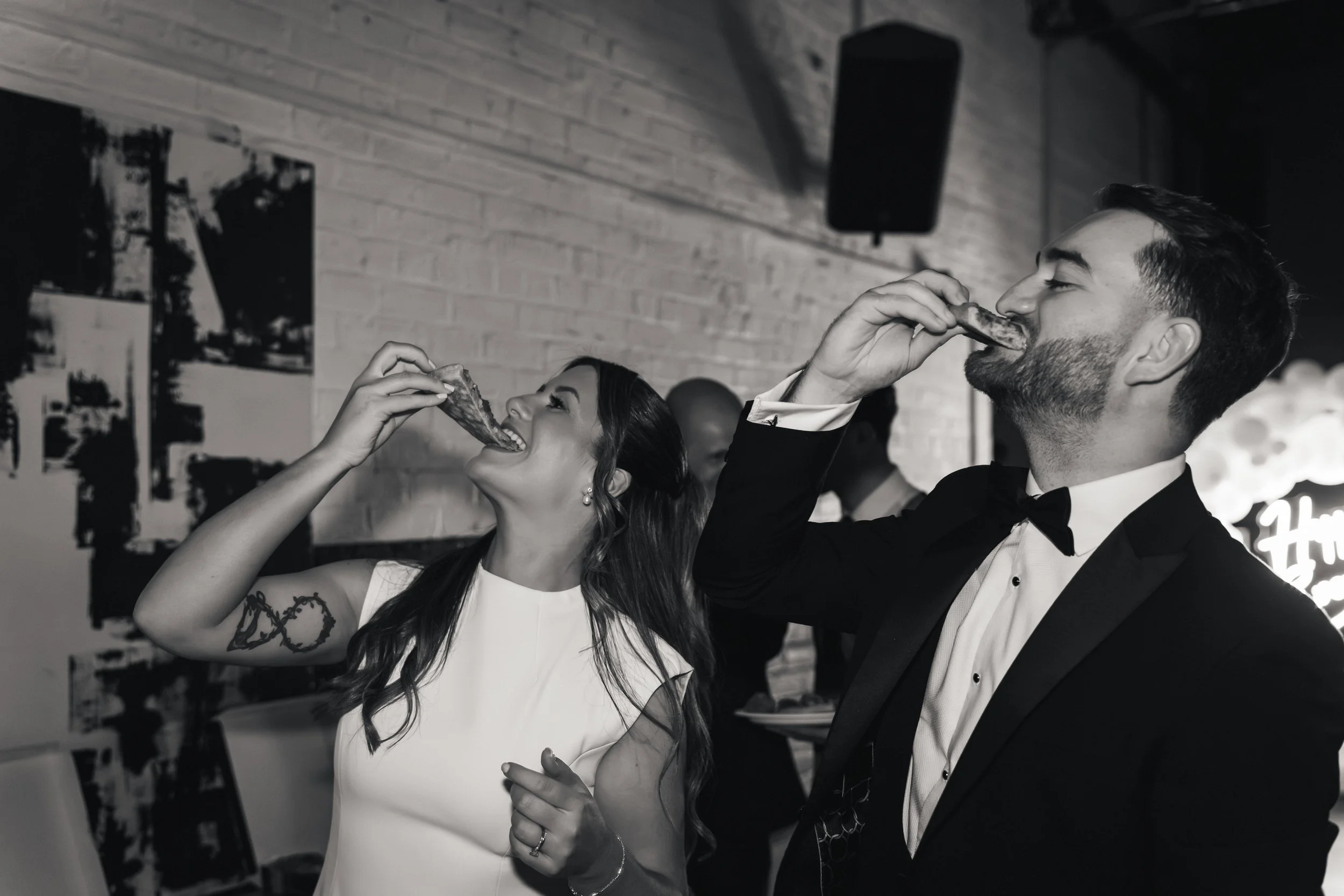 A man and woman in formal attire enjoying food at a celebration, both holding pieces of meat near their mouths for a bite.