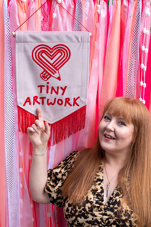 A woman with long red hair and a leopard print top points to a wall hanging that says 'Tiny Artwork' with a heart fishing wire design. The background is decorated with pink and red ribbons and lights.