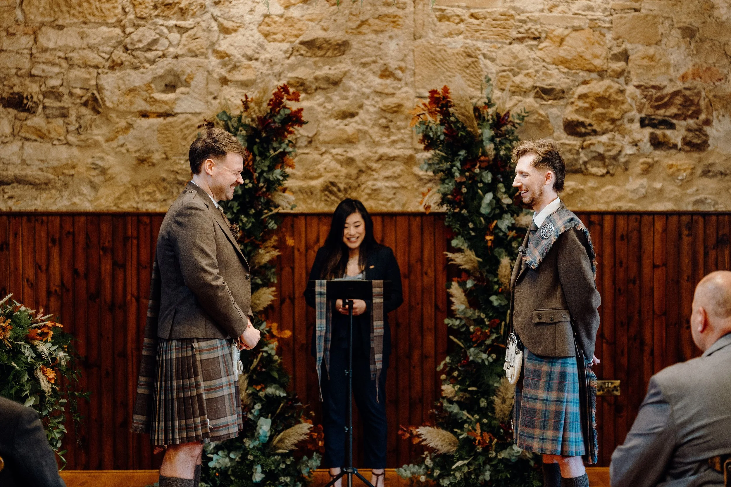 Two men in kilts facing each other and smiling during a wedding ceremony, with a woman reading vows in the background, decorated with floral arrangements and a stone wall.