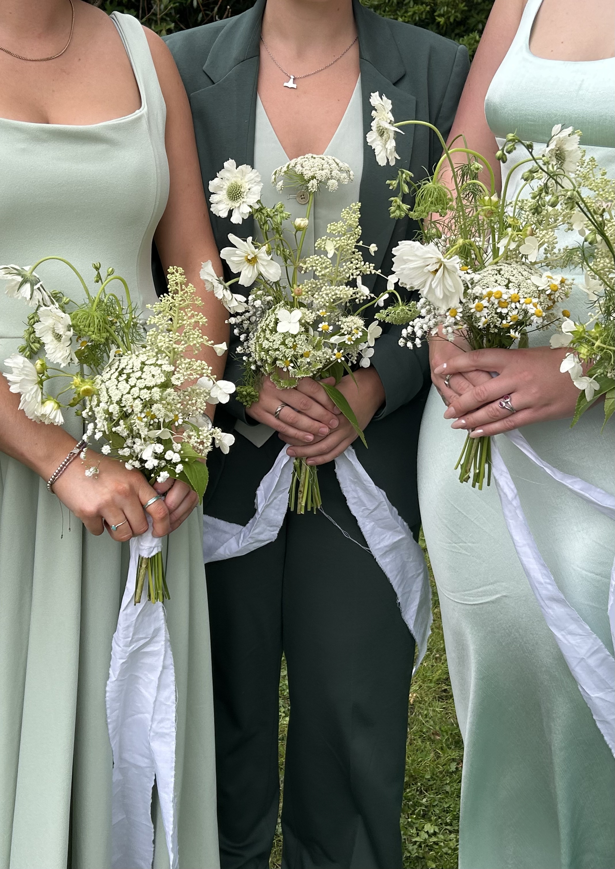 Three women in pastel dresses and a dark green suit holding bouquets of white flowers, standing outdoors on grass.