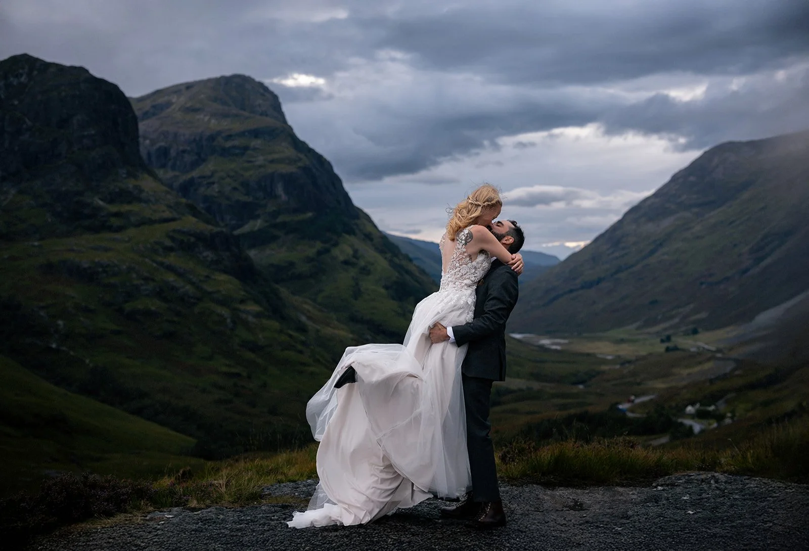 A couple in wedding attire embracing in a mountainous outdoor setting, with cloudy sky overhead.