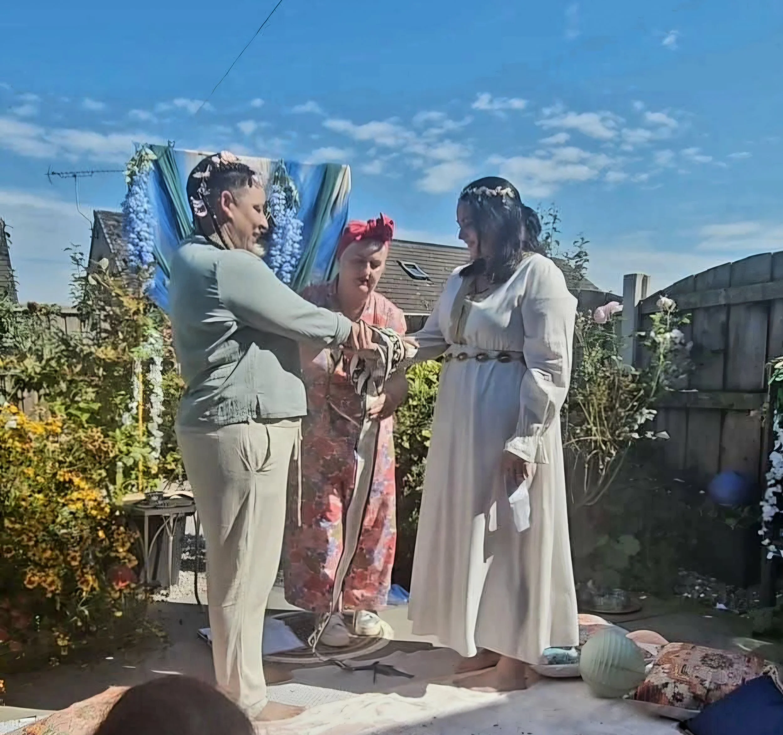 Three women participating in a wedding ceremony outdoors, with a blue sky and a garden fence in the background.