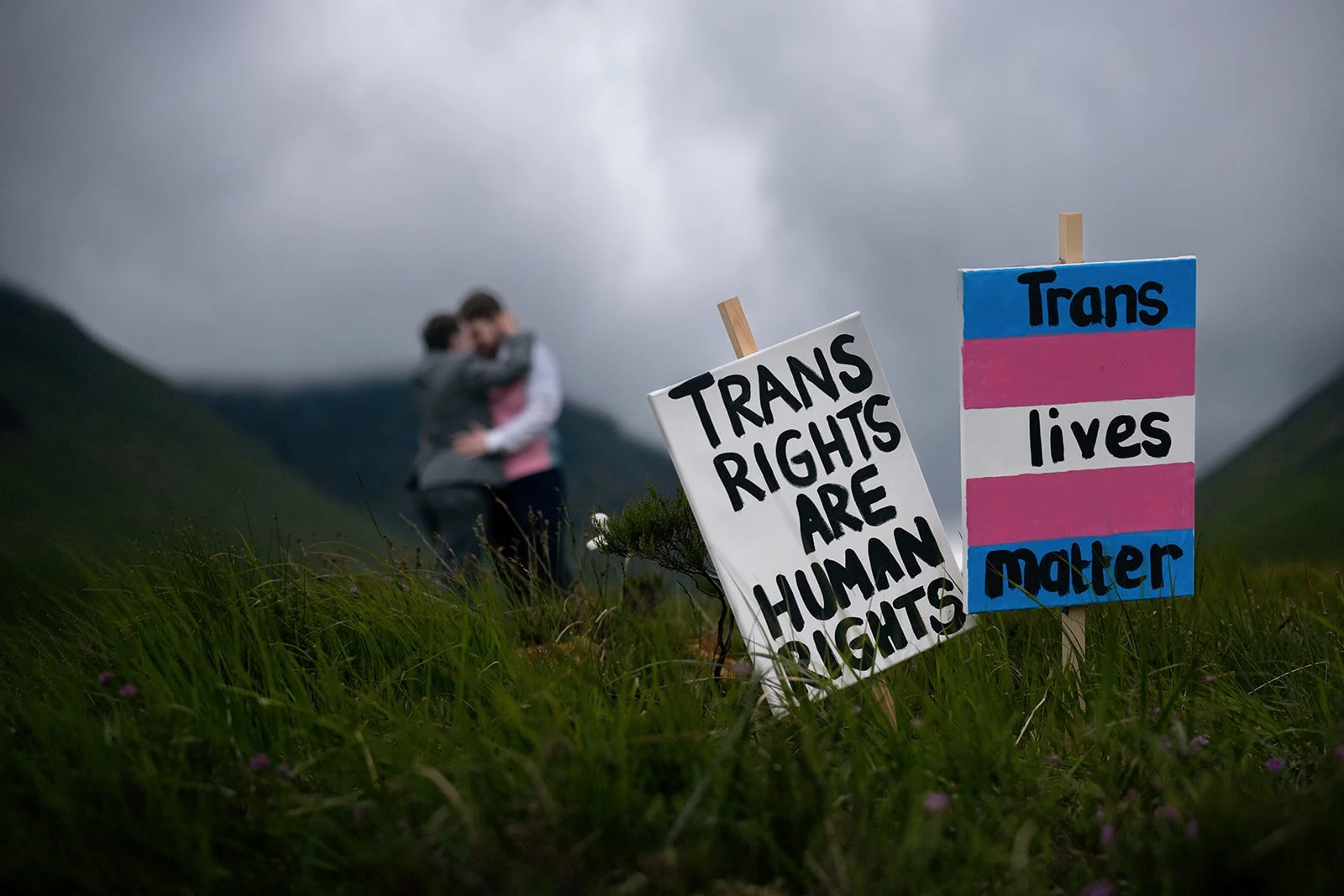 Two people embracing in a grassy outdoor area with mountains and cloudy sky in the background, and two signs on the ground. One sign reads "Trans rights are human rights" in black letters, and another sign with a transgender pride flag pattern reads "Trans lives matter" in black letters.