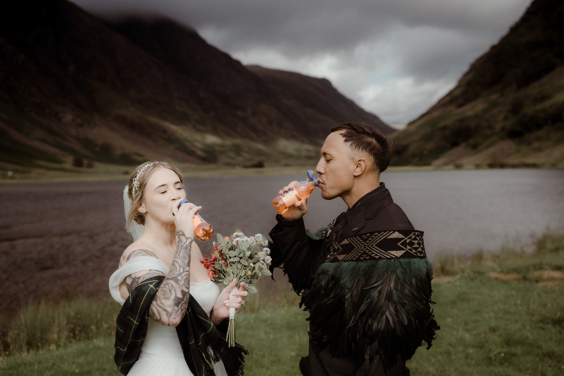 A bride and groom standing outdoors by a lake in a mountainous area, both drinking from water bottles, with the bride holding a bouquet of flowers, during cloudy weather.