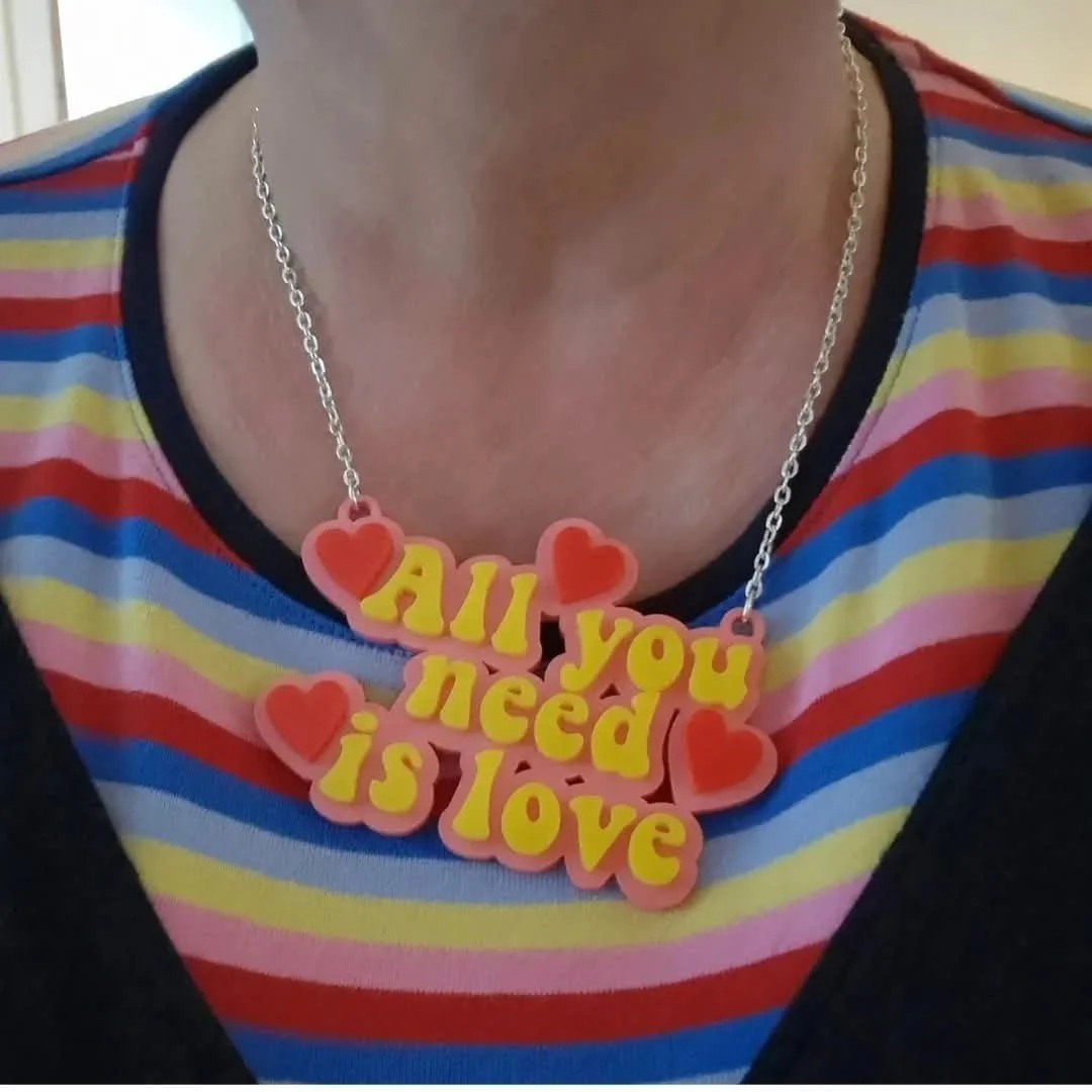 A person wearing a colorful striped shirt with a necklace that reads "All you need is love" and has red hearts around the text.