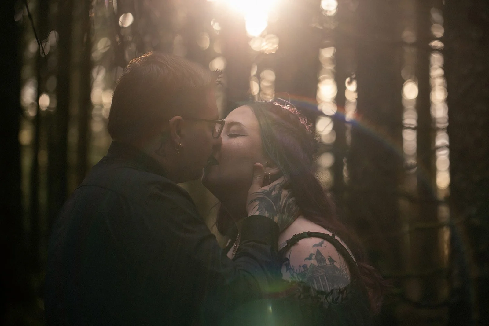 A couple sharing an intimate kiss at sunset, with backlighting creating a romantic atmosphere, in a wooded area with trees.