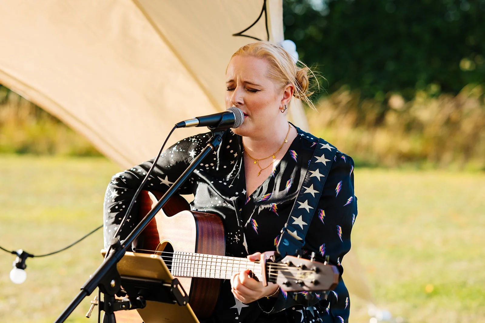 A woman with blonde hair singing into a microphone while playing an acoustic guitar outdoors, under a large tent with trees in the background.