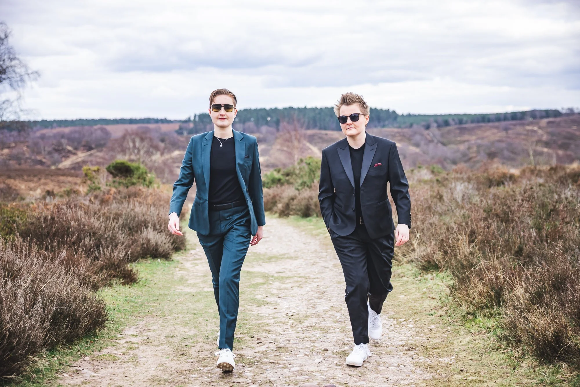 Two women in suits walking on a dirt path in a rural outdoor landscape with cloudy sky