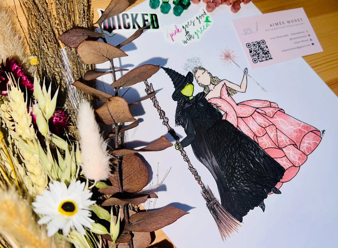 A colorful hand-drawn illustration of two women, one dressed as a witch with a pointed hat and green face, and the other wearing a pink dress with a star-shaped wand, sitting on a broomstick. The illustration is on a white sheet of paper, surrounded by dried and fresh flowers, and a business card is in the upper right corner.