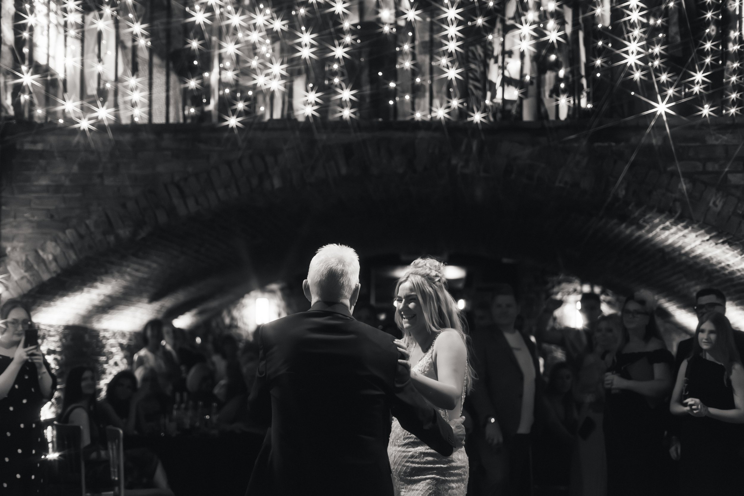A black and white photo of a couple dancing at a celebration, with people watching in the background and star-shaped lights above.