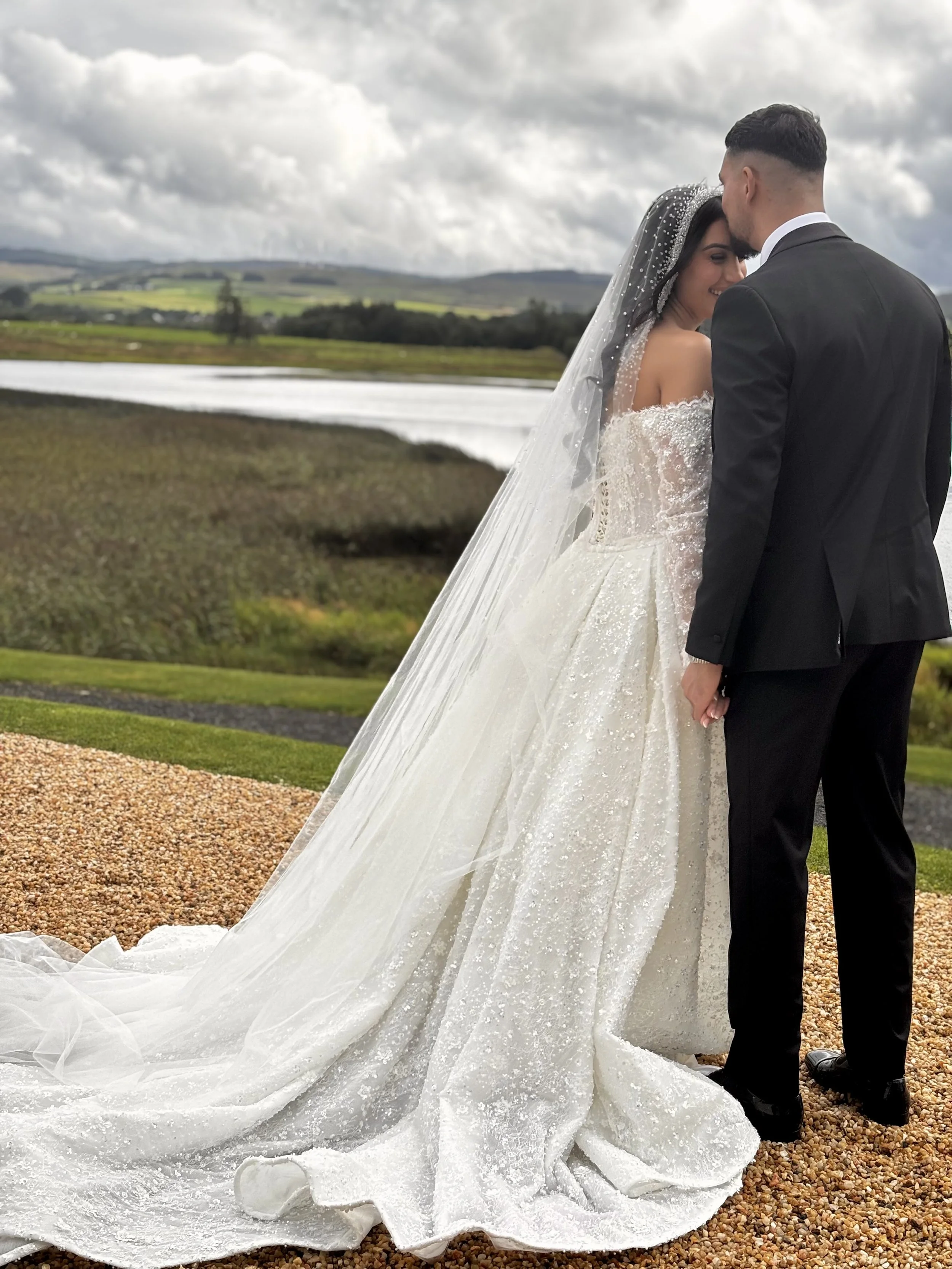 A bride and groom standing close outdoors on a gravel path by a body of water, with rolling hills and cloudy sky in the background; the bride wears a white wedding gown with a long veil and the groom wears a black suit.