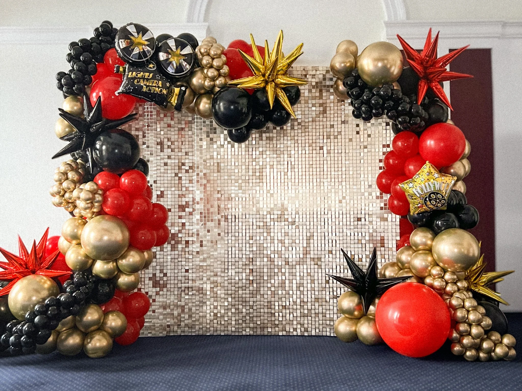 Decorative balloon arrangement featuring black, red, and gold balloons of various sizes and shapes, including star-shaped and round balloons, with a metallic mosaic background.