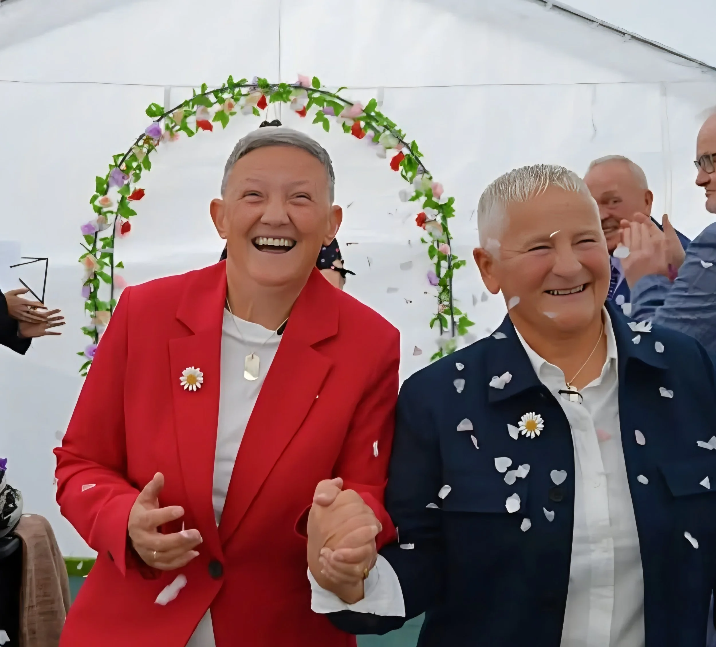 Two smiling people, a woman in a red blazer and a man in a dark jacket, are celebrating with confetti at a festive event. They are holding hands and appear joyful.