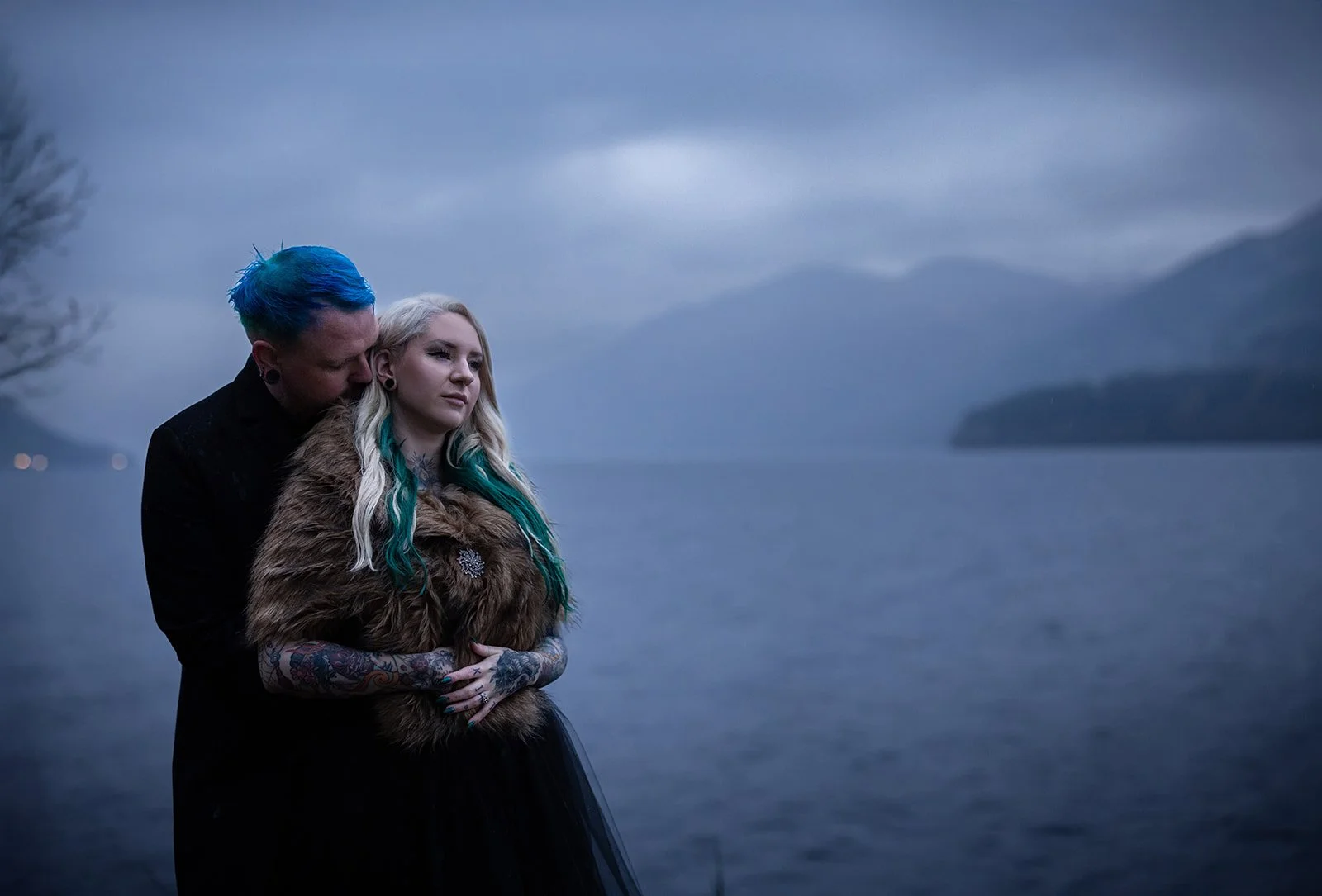A man with blue hair and a woman with blonde hair and teal highlights standing by a body of water with mountains in the background.