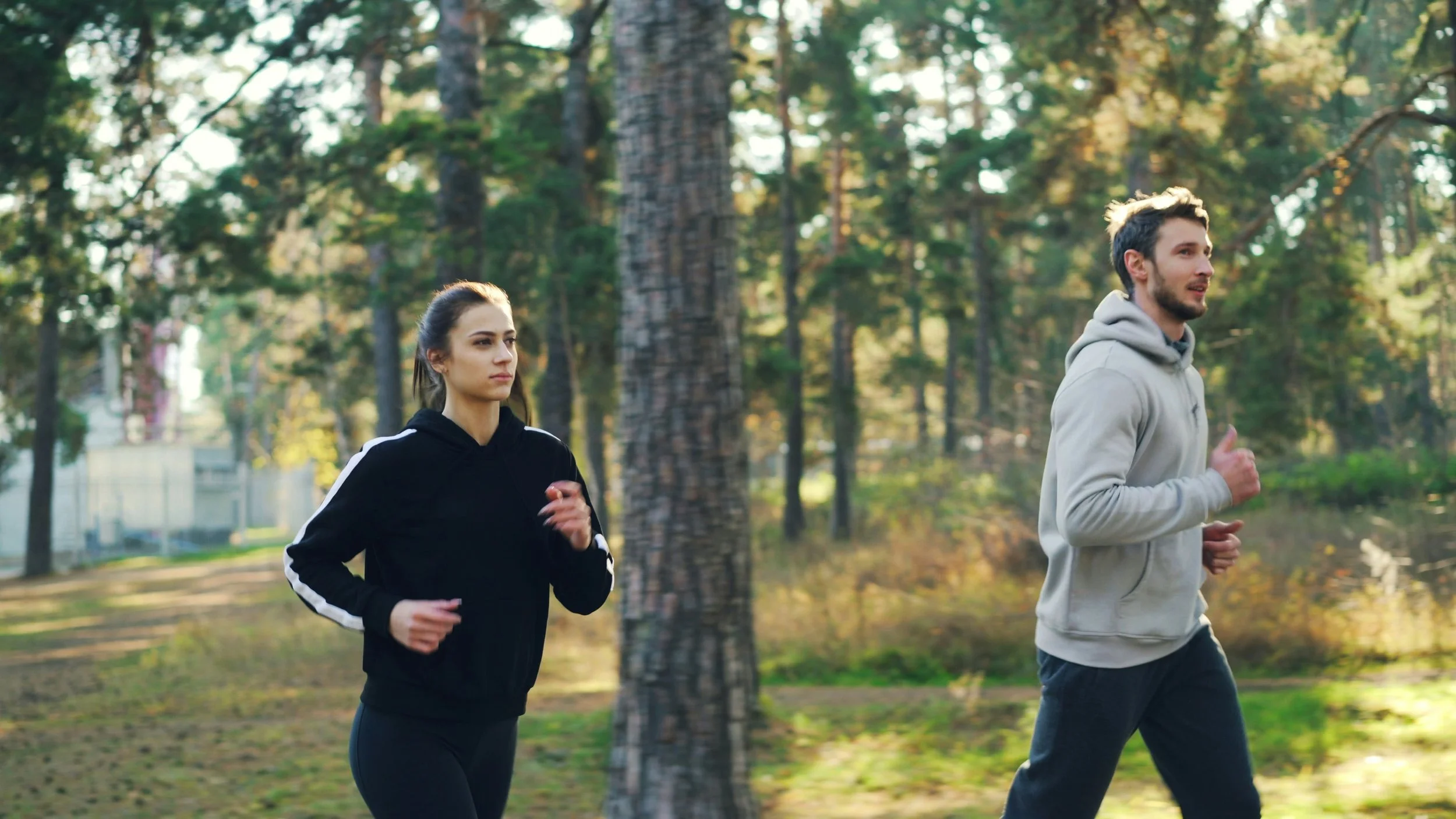 A man and woman jogging outdoors in a wooded park, both dressed in athletic clothing.