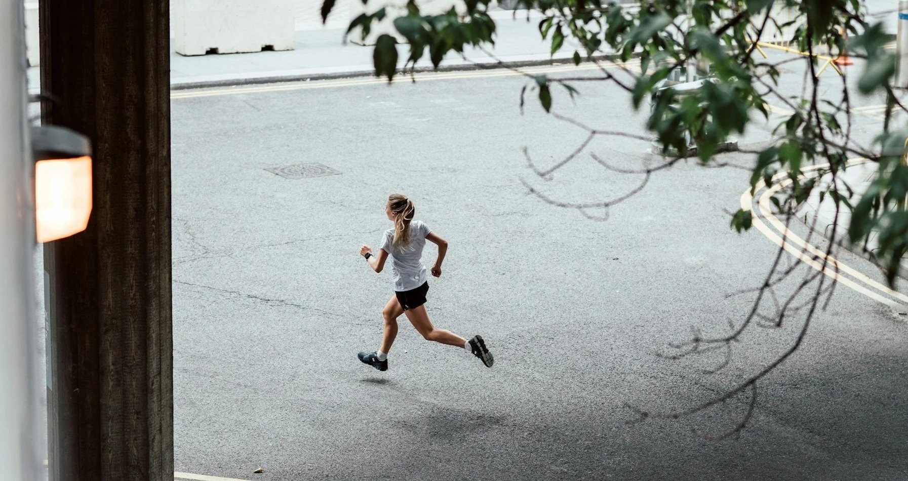 A woman jogging on an empty street seen from a high angle, with branches and leaves partially framing the top of the image.