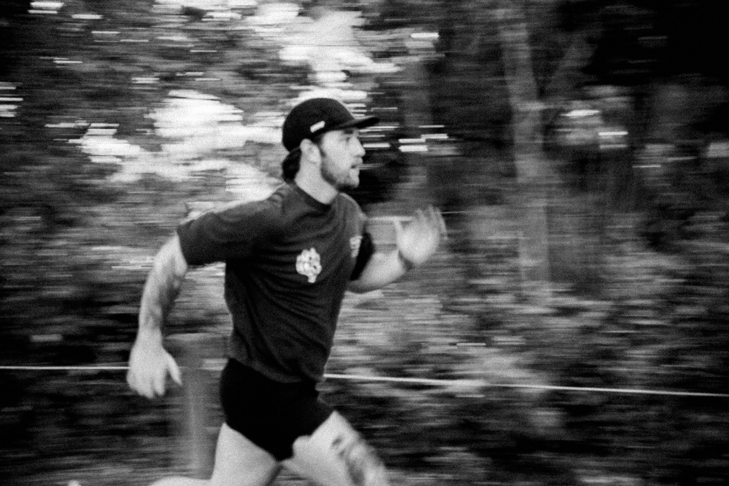 A man running outdoors in motion blur, wearing a black cap, black shirt, and shorts, with trees in the background.