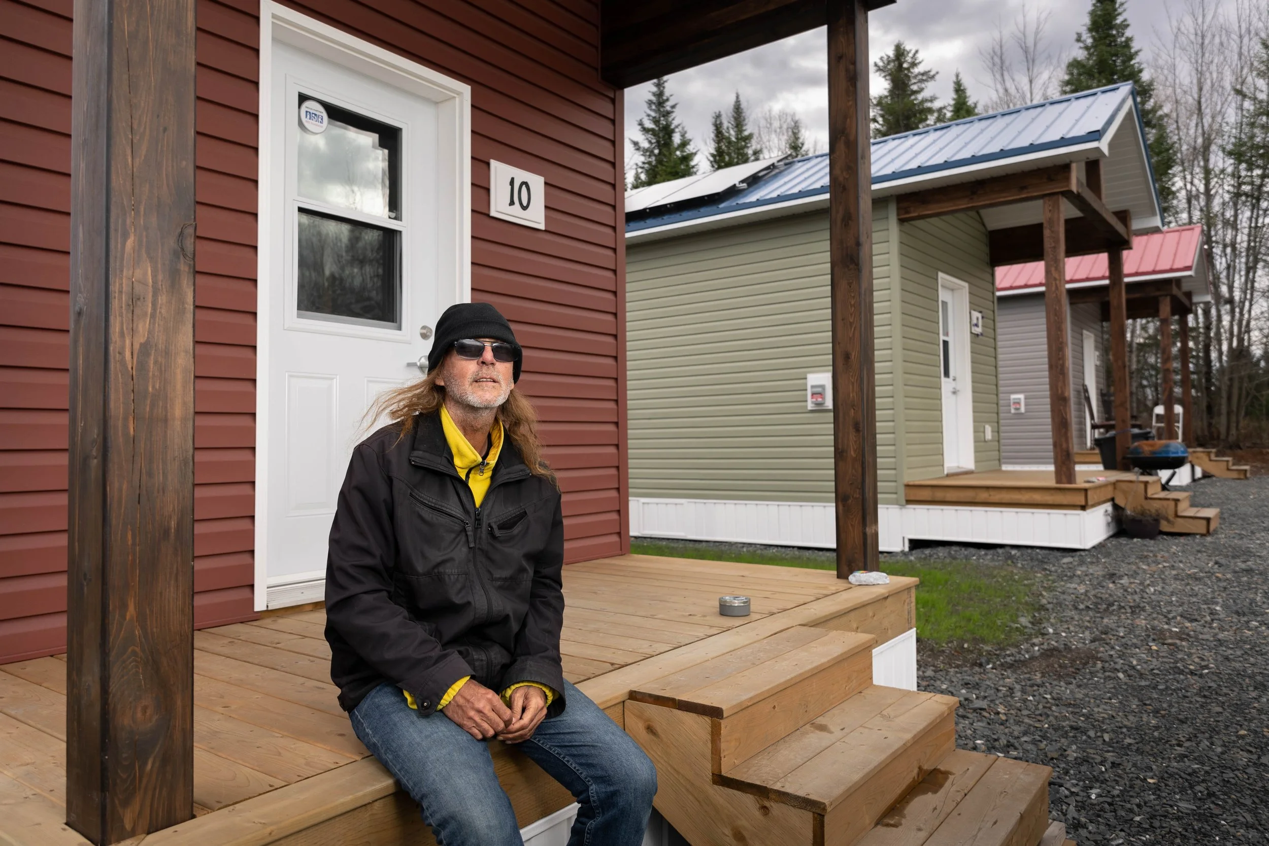 A man with long hair, a black beanie, glasses, a yellow shirt, a black jacket, and blue jeans sitting on a wooden porch in front of a reddish-brown house with a white door, with other similar houses painted in grey and green in the background, during overcast weather.