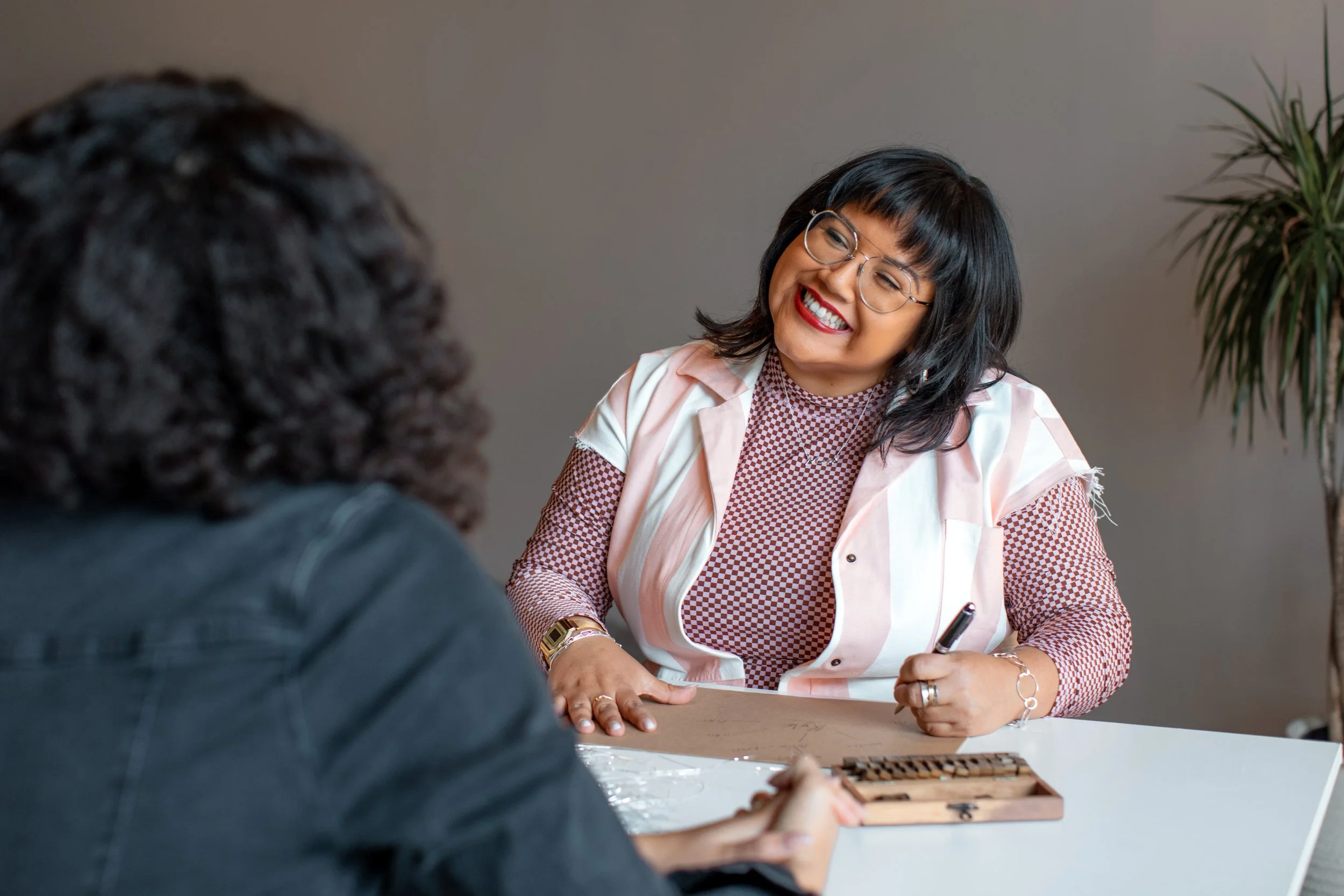 A woman with black hair, glasses, and red lipstick smiling, sitting at a table during a conversation with another person.