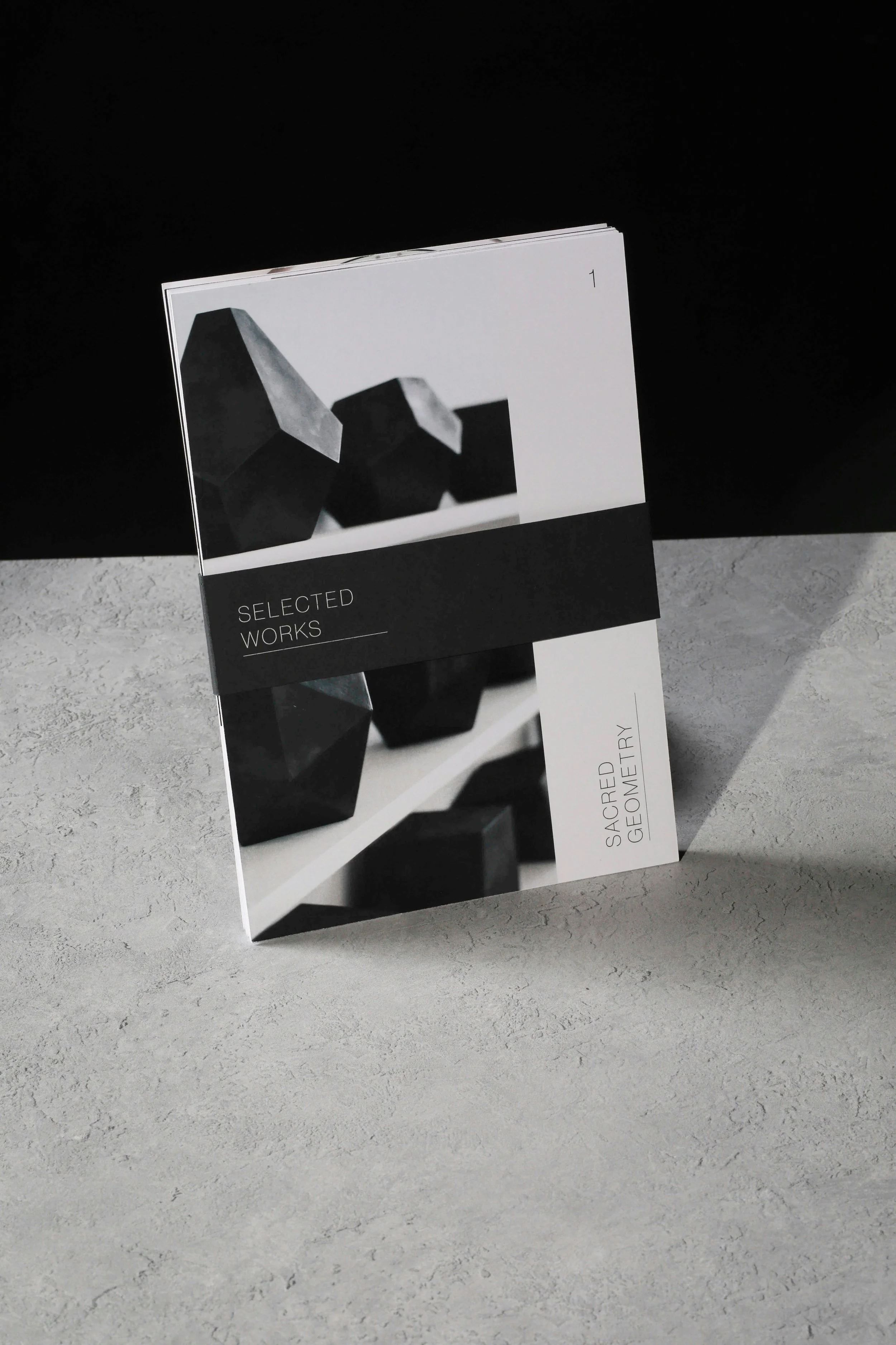 A black and white book titled "Selected Works" with the subtitle "Sacred Geometries" stands upright on a textured light-colored surface against a dark background.